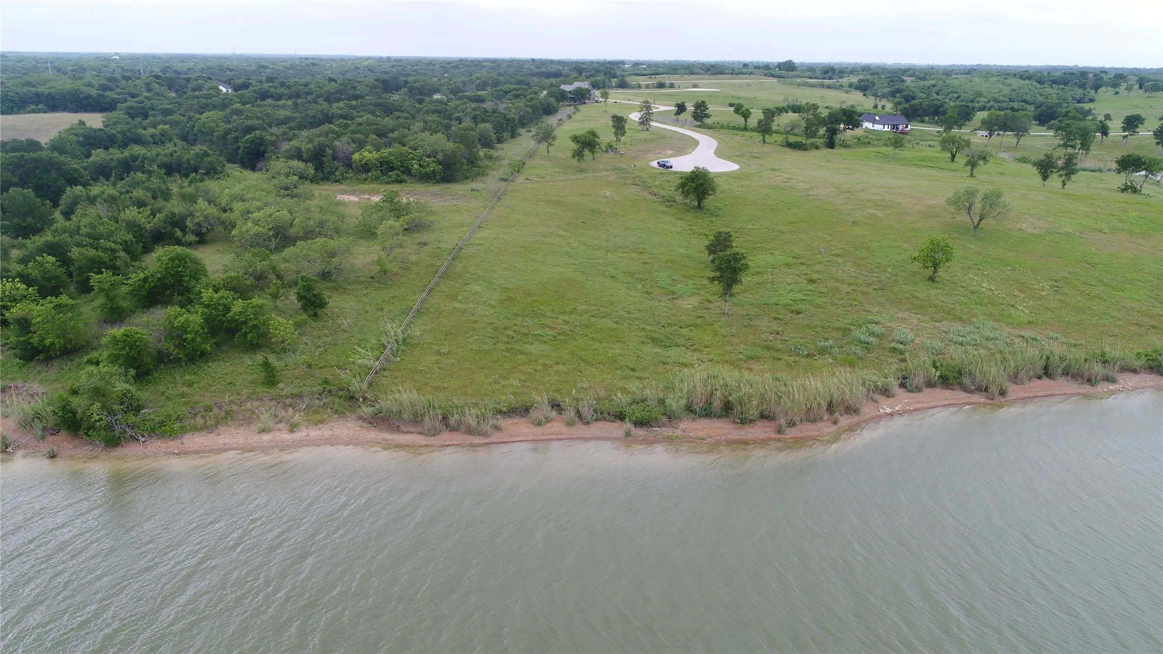 Expansive waterfront property featuring a natural shoreline, mature trees, open grassy areas, and a paved cul-de-sac