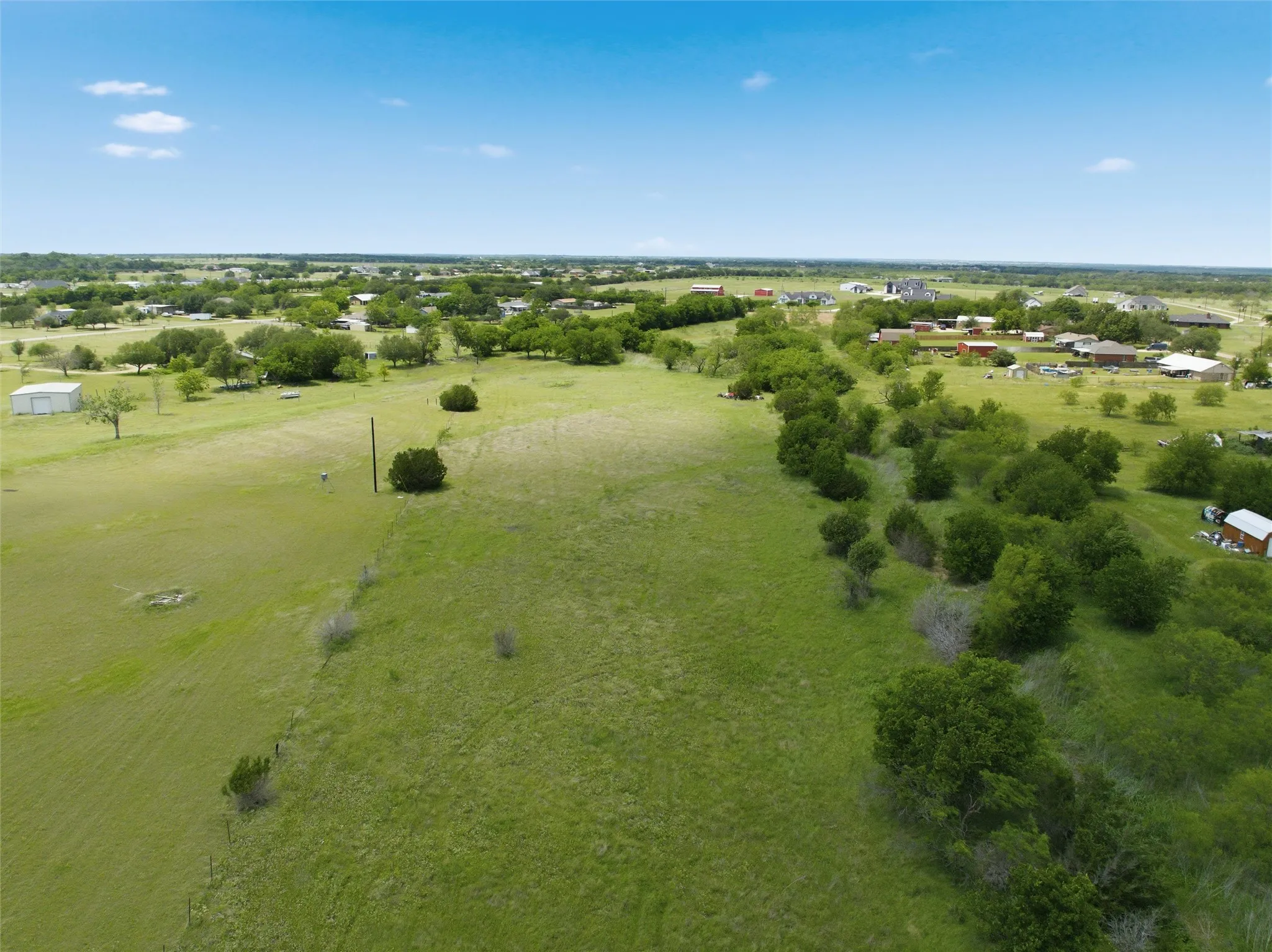 Expansive green acreage with scattered mature trees and distant residential structures under a clear sky