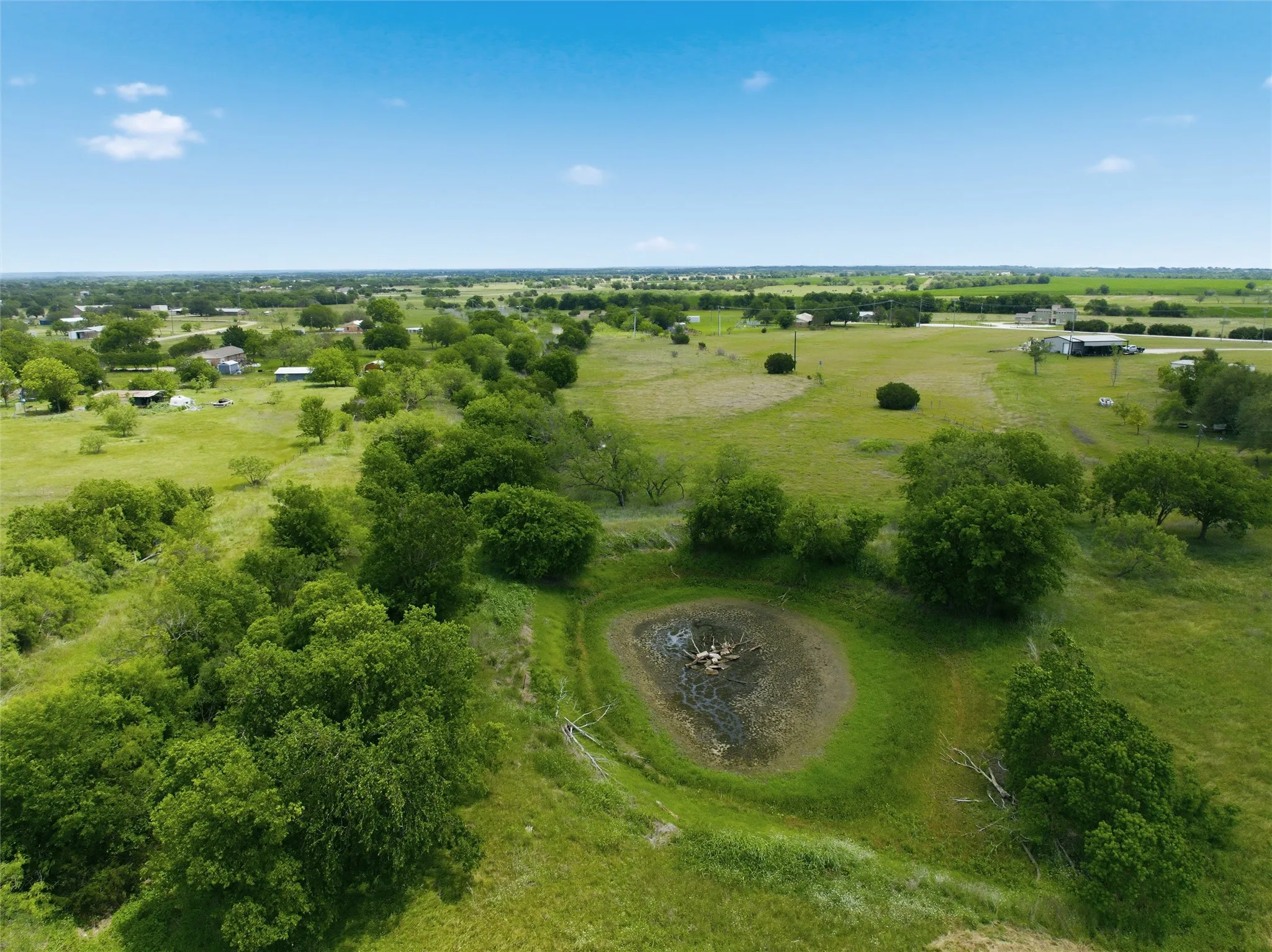 Expansive green landscape featuring a natural pond, scattered mature trees, open fields, and distant residential structures