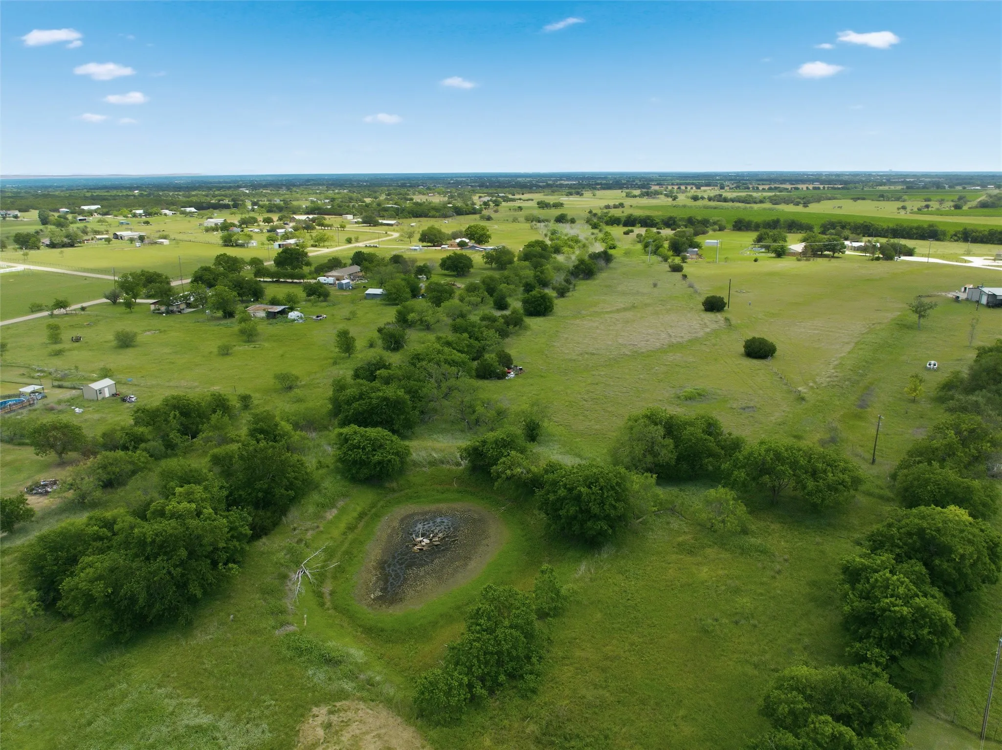 Expansive green acreage featuring a natural pond, mature trees, and open fields under a blue sky