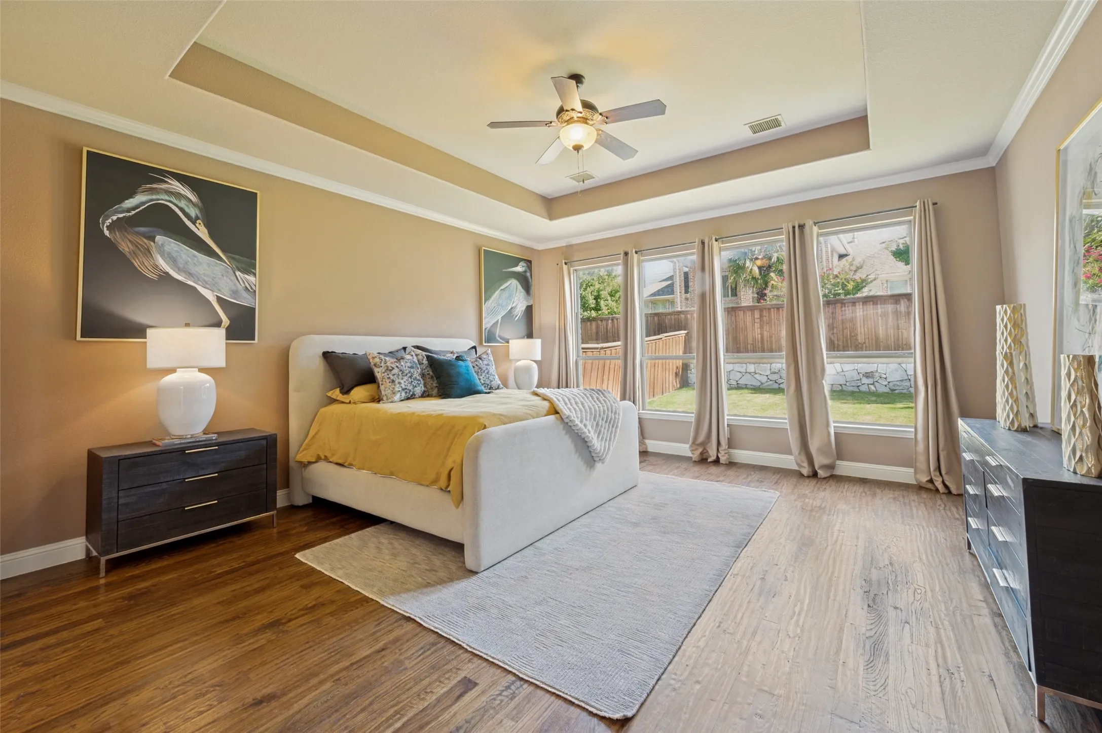 Spacious primary suite designed as a private retreat, featuring elegant tray ceilings, rich hardwood flooring, and a wall of windows that fill the room with natural light while overlooking the backyard. Generous proportions provide plenty of space for a sitting area, creating the perfect place to relax and unwind.