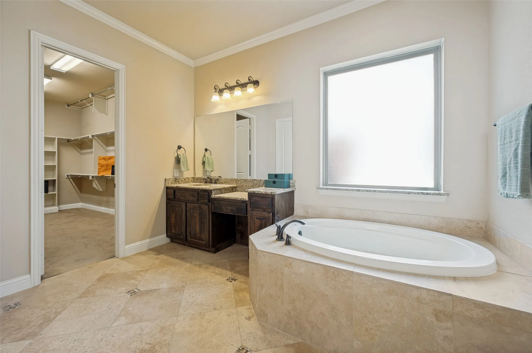 Spa-inspired primary ensuite featuring a relaxing garden tub beneath a large privacy window, a built-in vanity area, and elegant natural stone finishes. The spacious layout flows seamlessly into the oversized walk-in closet, creating a luxurious and functional owner's retreat.