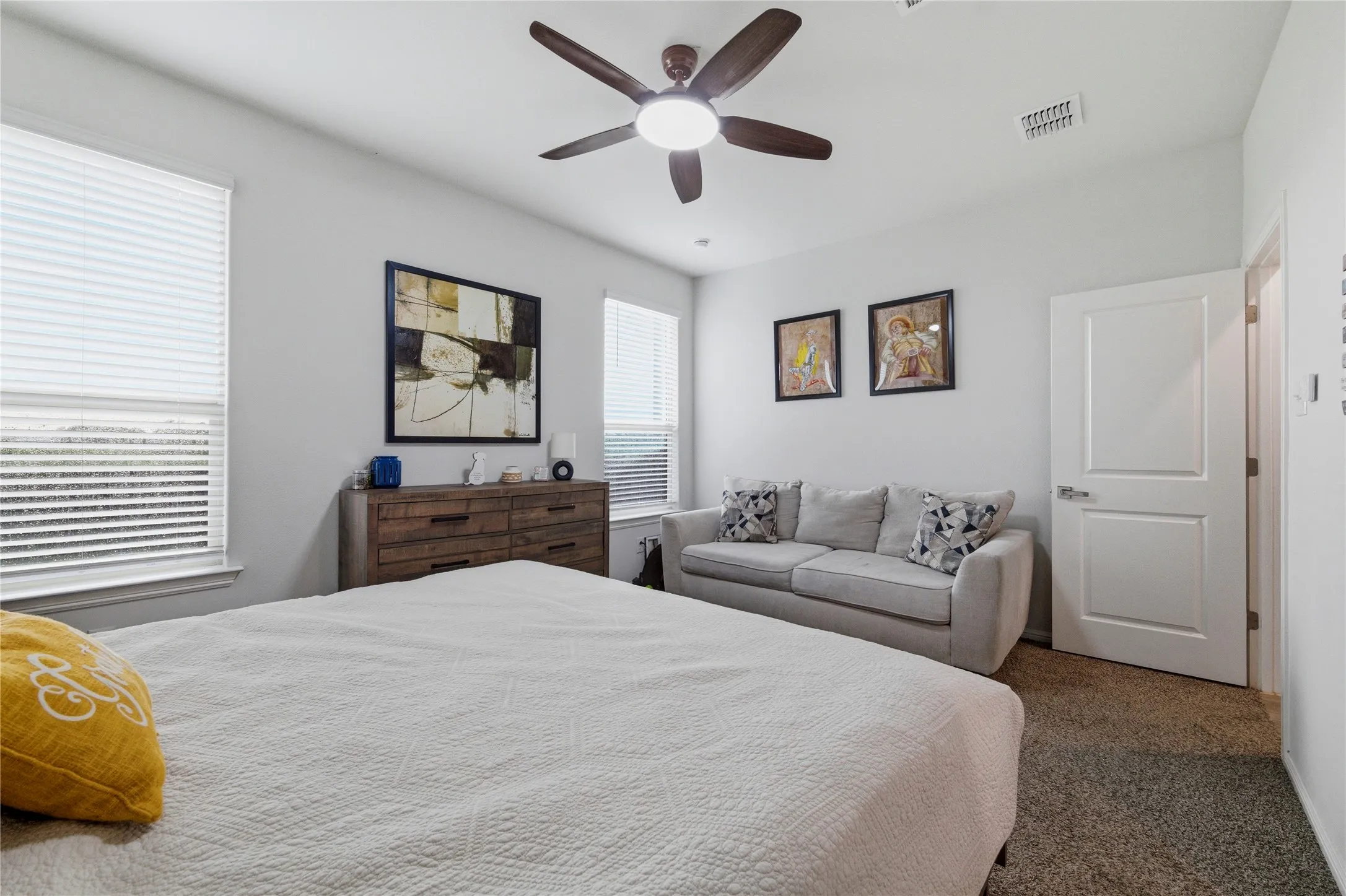 Spacious bedroom featuring light neutral wall paint, two windows with blinds, a ceiling fan with integrated lighting, and carpet flooring