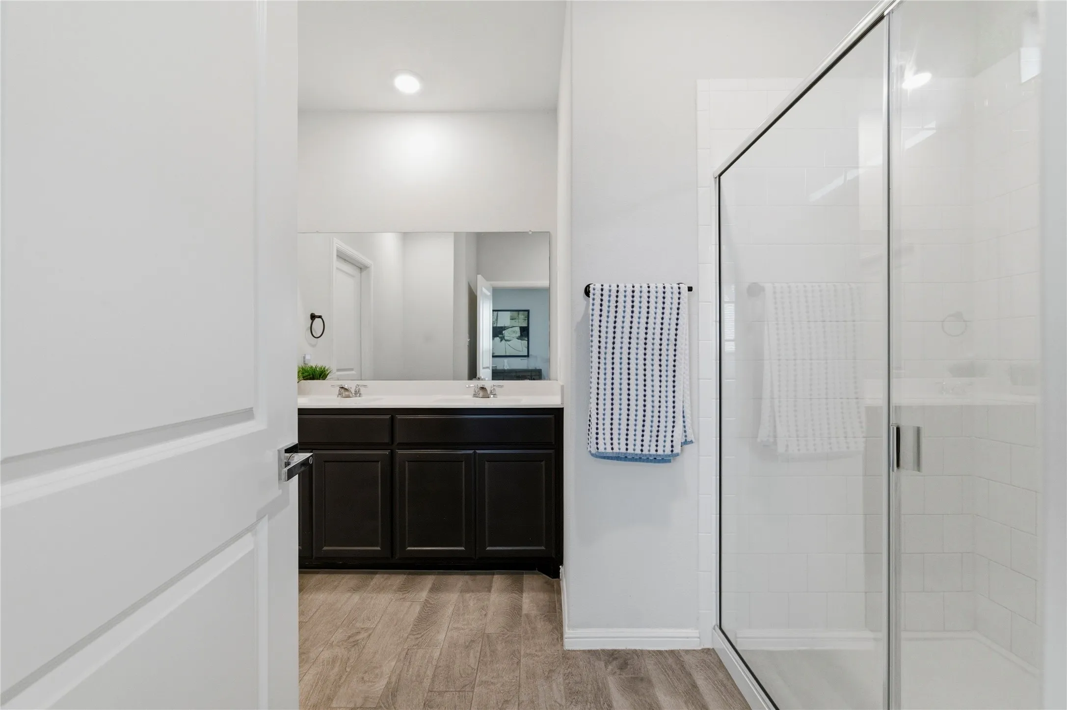 Bathroom featuring a glass-enclosed shower with white subway tile, a dual vanity with dark wood-finish cabinetry, a large mirror, and wood-finish flooring