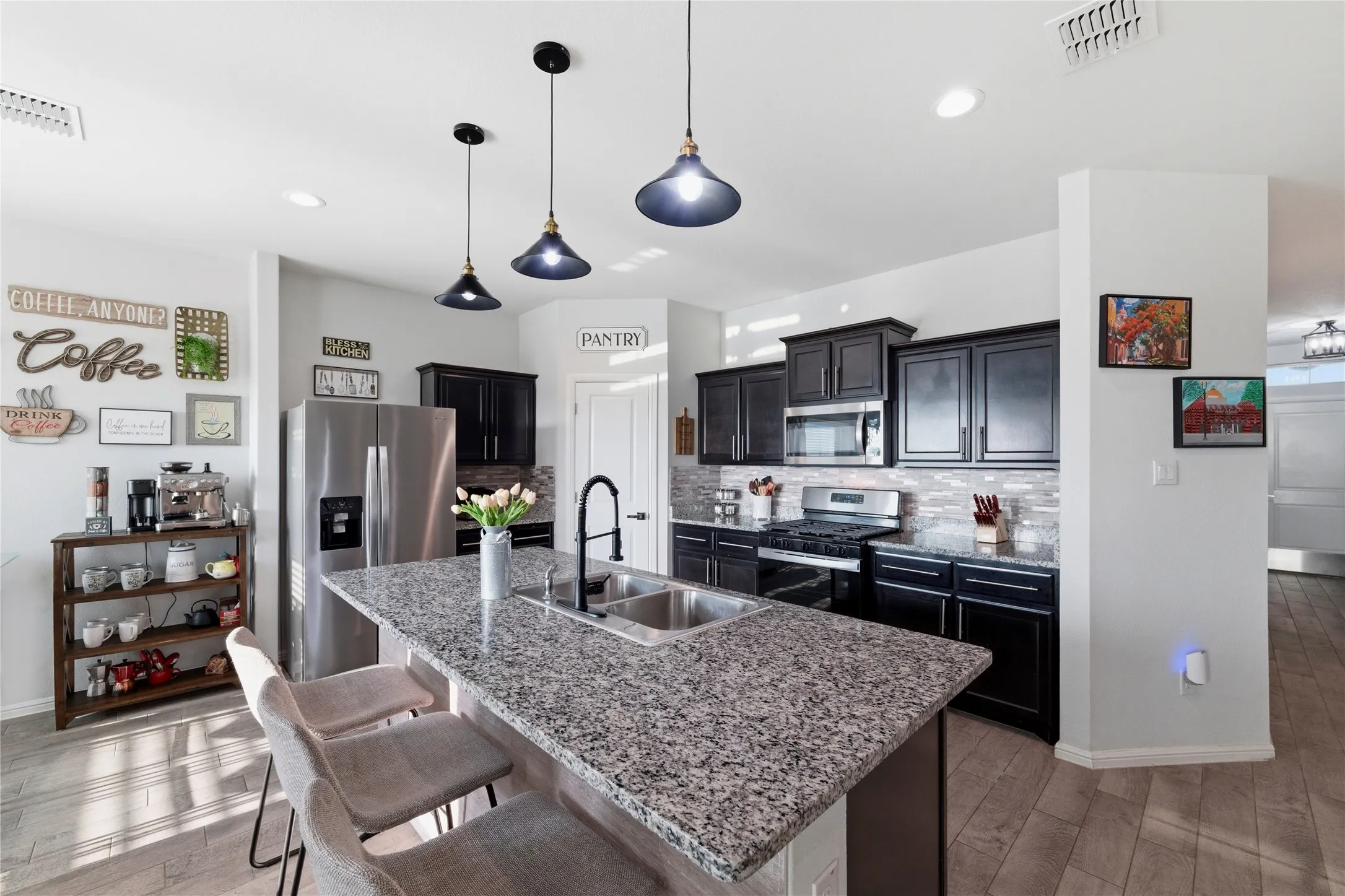Kitchen featuring a granite-topped island with an undermount double sink, dark wood cabinetry, stainless steel appliances, a tiled backsplash, and pendant lighting