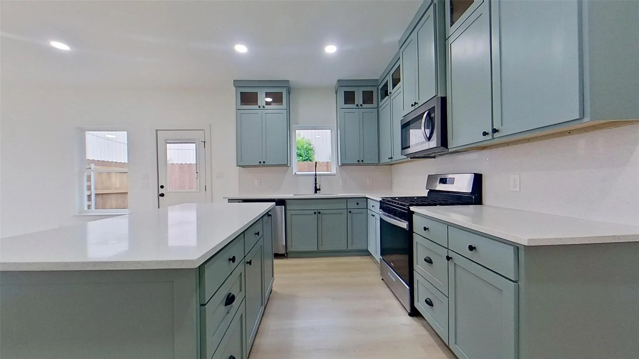 Spacious kitchen featuring an expansive central island with a solid surface countertop, light-toned wood-finish flooring, abundant cabinetry with black hardware, white subway tile backsplash, and stainless steel appliances including a microwave and range