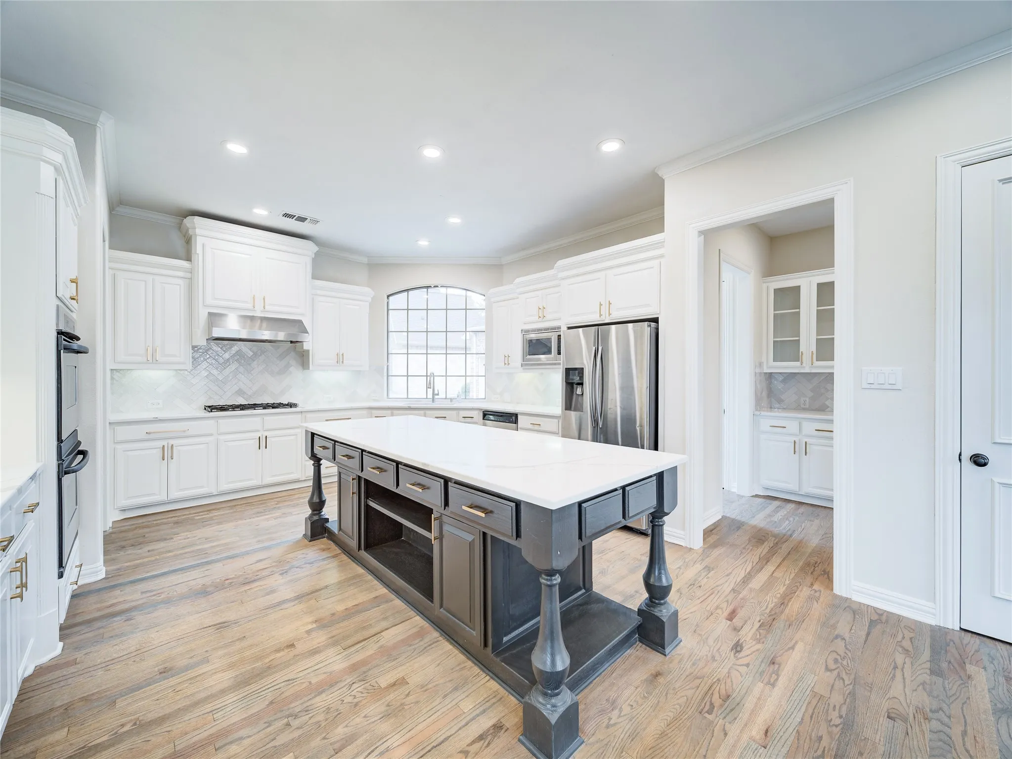 Gourmet kitchen featuring a large central island with a solid surface countertop, wood-finish flooring, white cabinetry with gold-tone hardware, stainless steel appliances, and a tiled backsplash