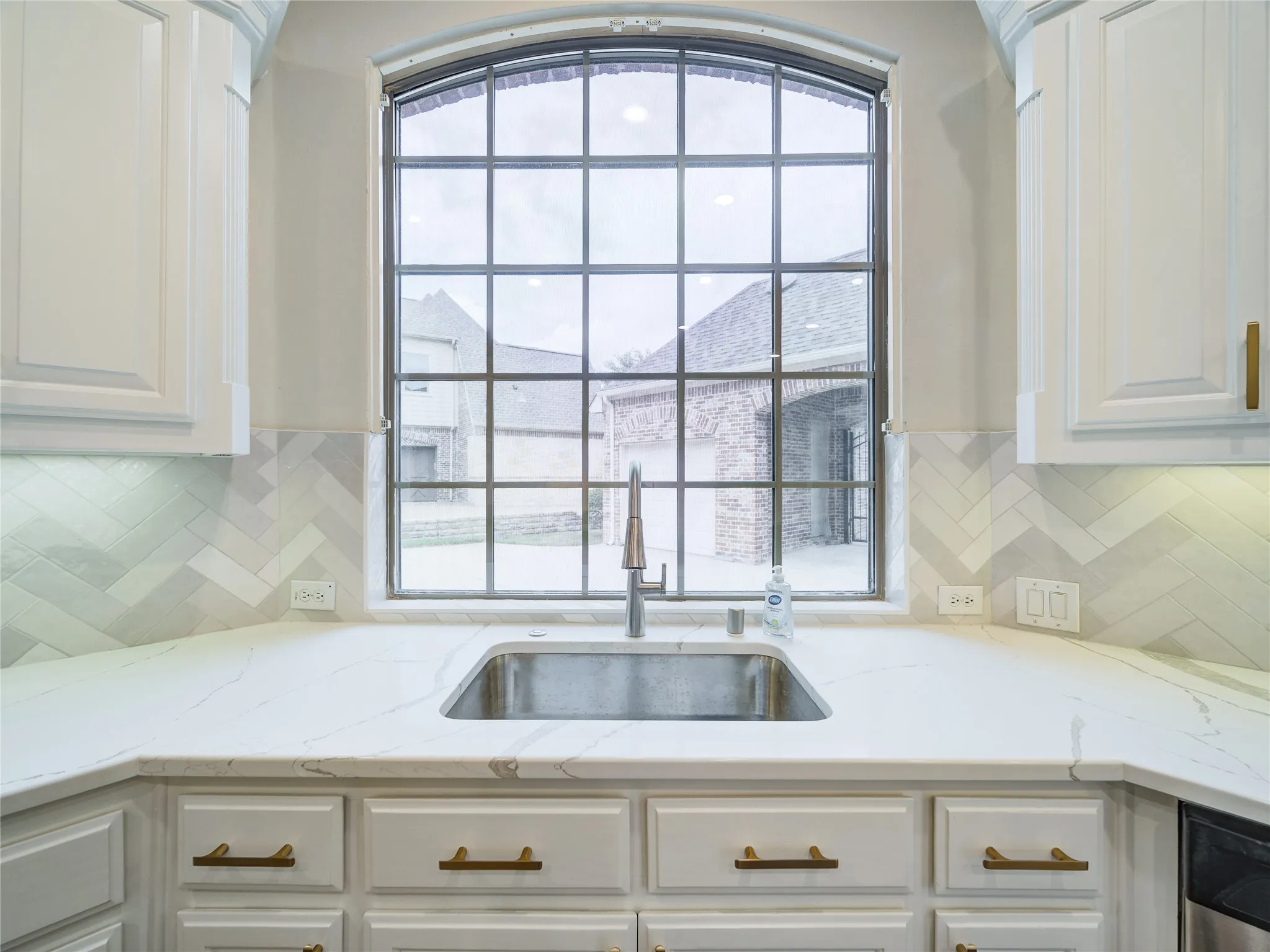 Kitchen sink area featuring an arched window, white cabinetry with brass hardware, a stainless steel sink, a modern faucet, and a herringbone tile backsplash