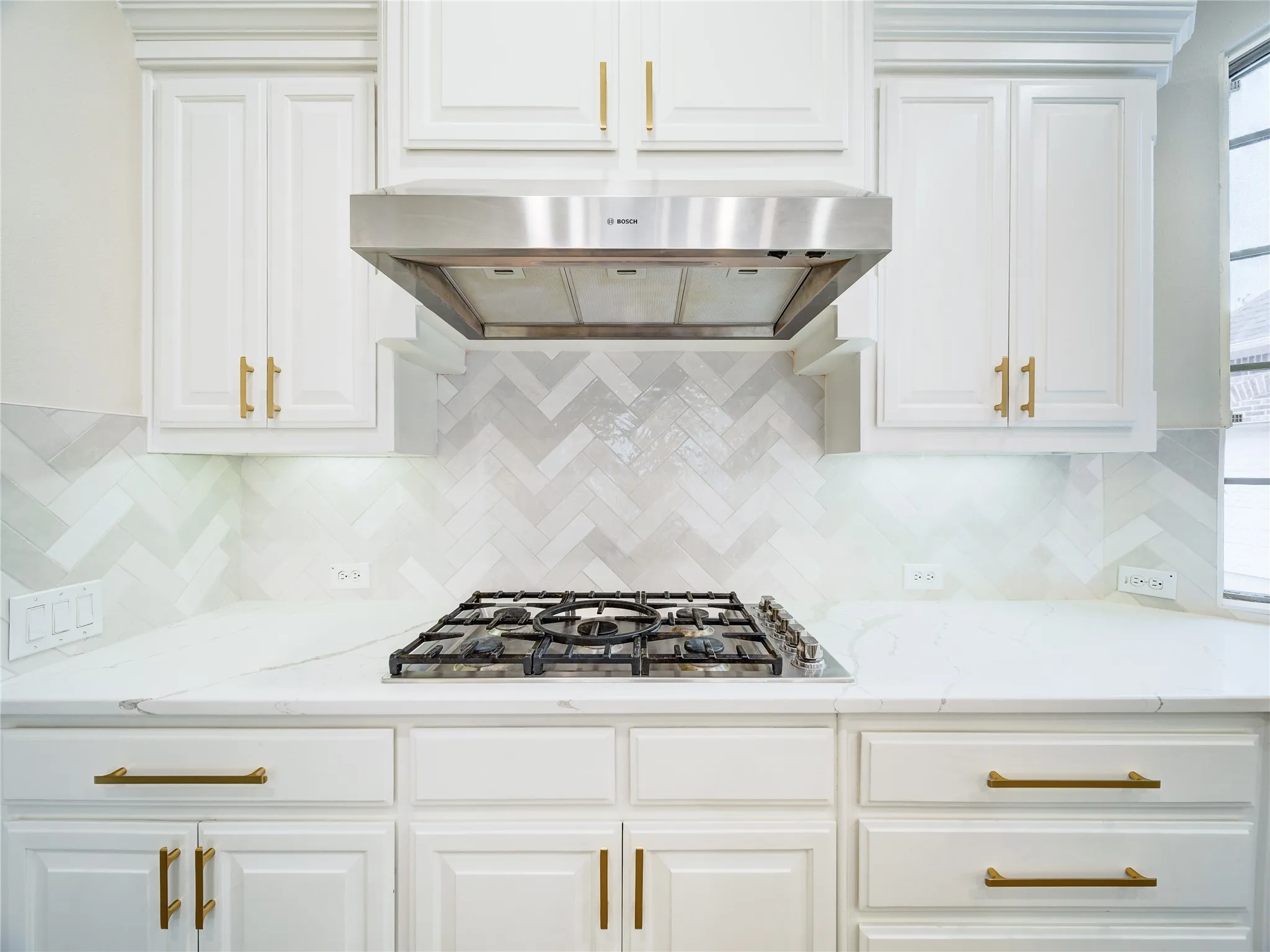 Kitchen featuring white cabinetry with gold-toned hardware, a stainless steel range hood, a gas cooktop, white countertops, and a herringbone pattern tile backsplash