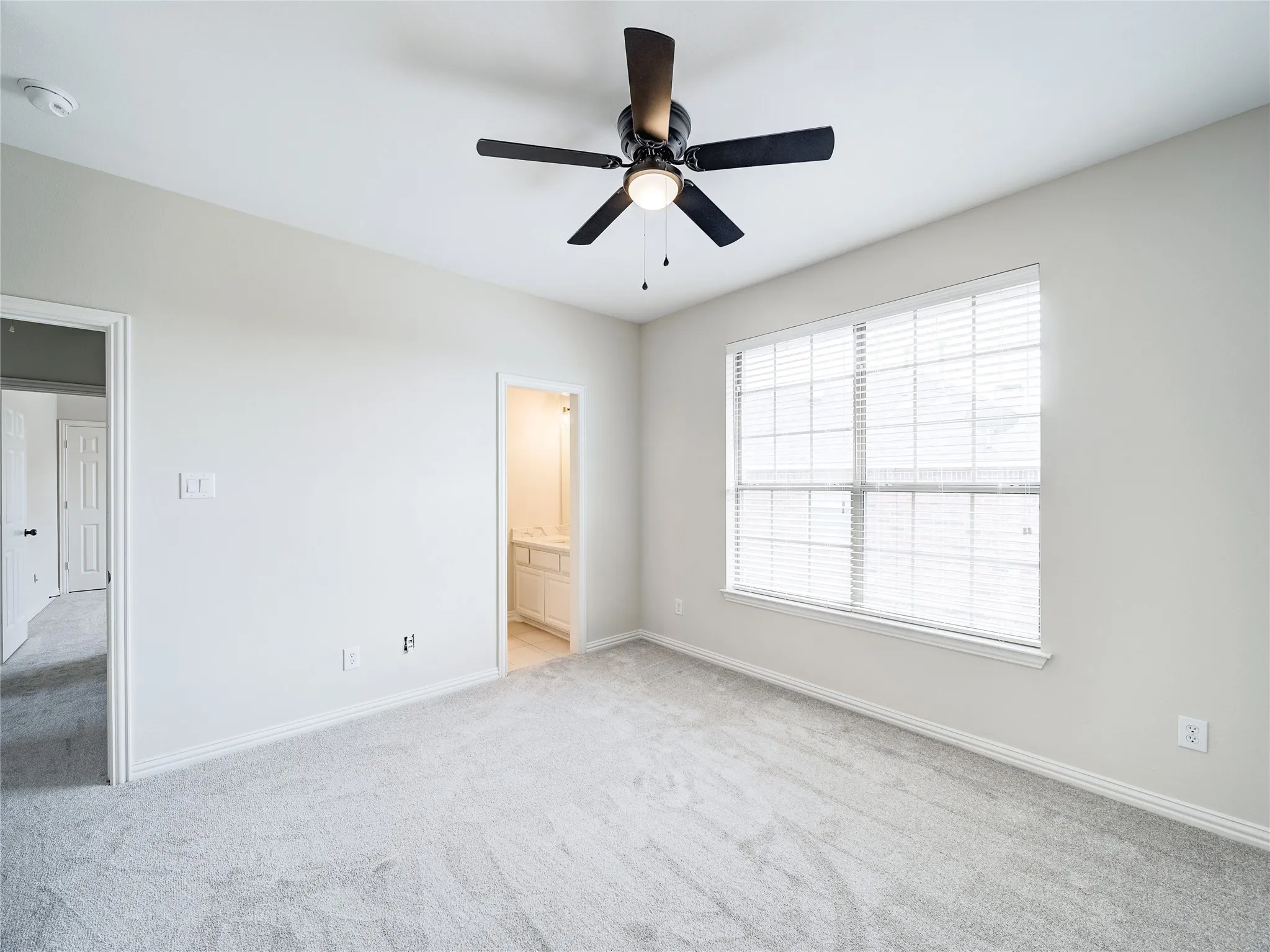 Carpeted room with a ceiling fan, a window with blinds, and an open doorway revealing a bathroom vanity