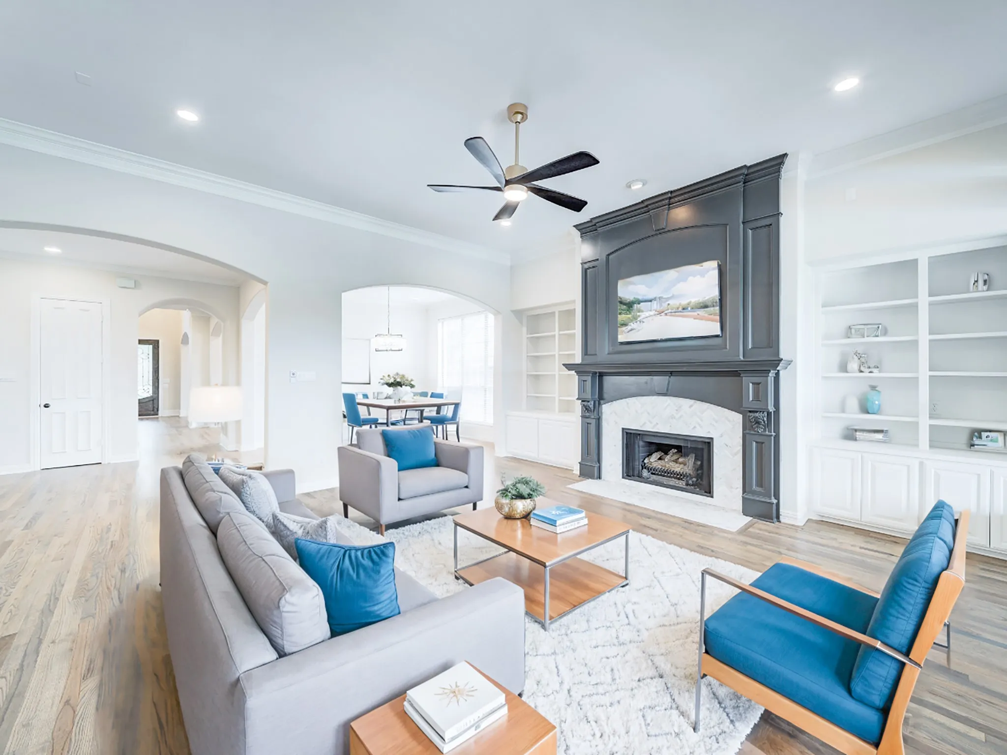 Spacious living area featuring light wood-finish flooring, a contemporary ceiling fan, and a fireplace with a dark-paneled surround and white herringbone tile