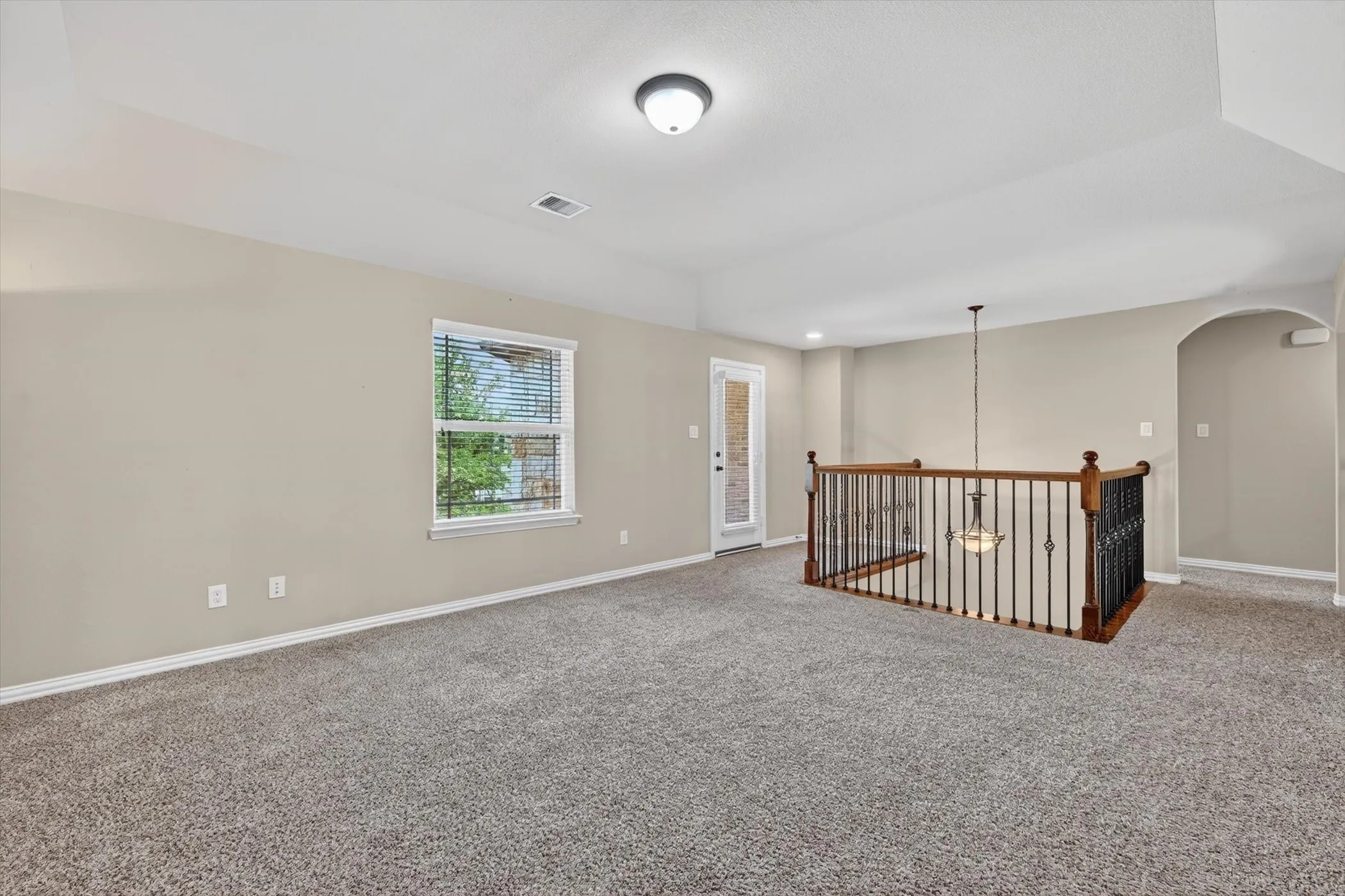 Carpeted loft area featuring a window with blinds, a glass-paneled door, and a staircase with wood and wrought iron railings