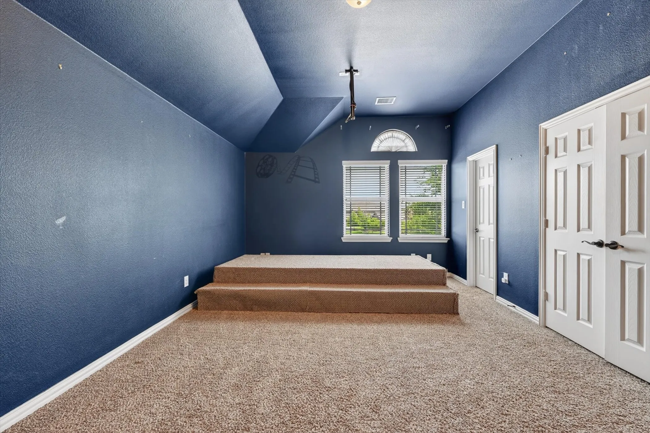 Carpeted room featuring a raised platform with two steps, two windows with blinds, an arched transom window, and two white paneled doors