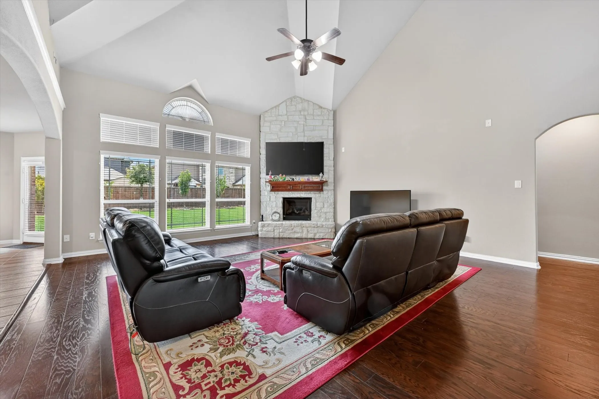 Spacious living area featuring vaulted ceilings, wood-finish flooring, a stone-faced fireplace with a wood mantel, and large windows with an arched transom