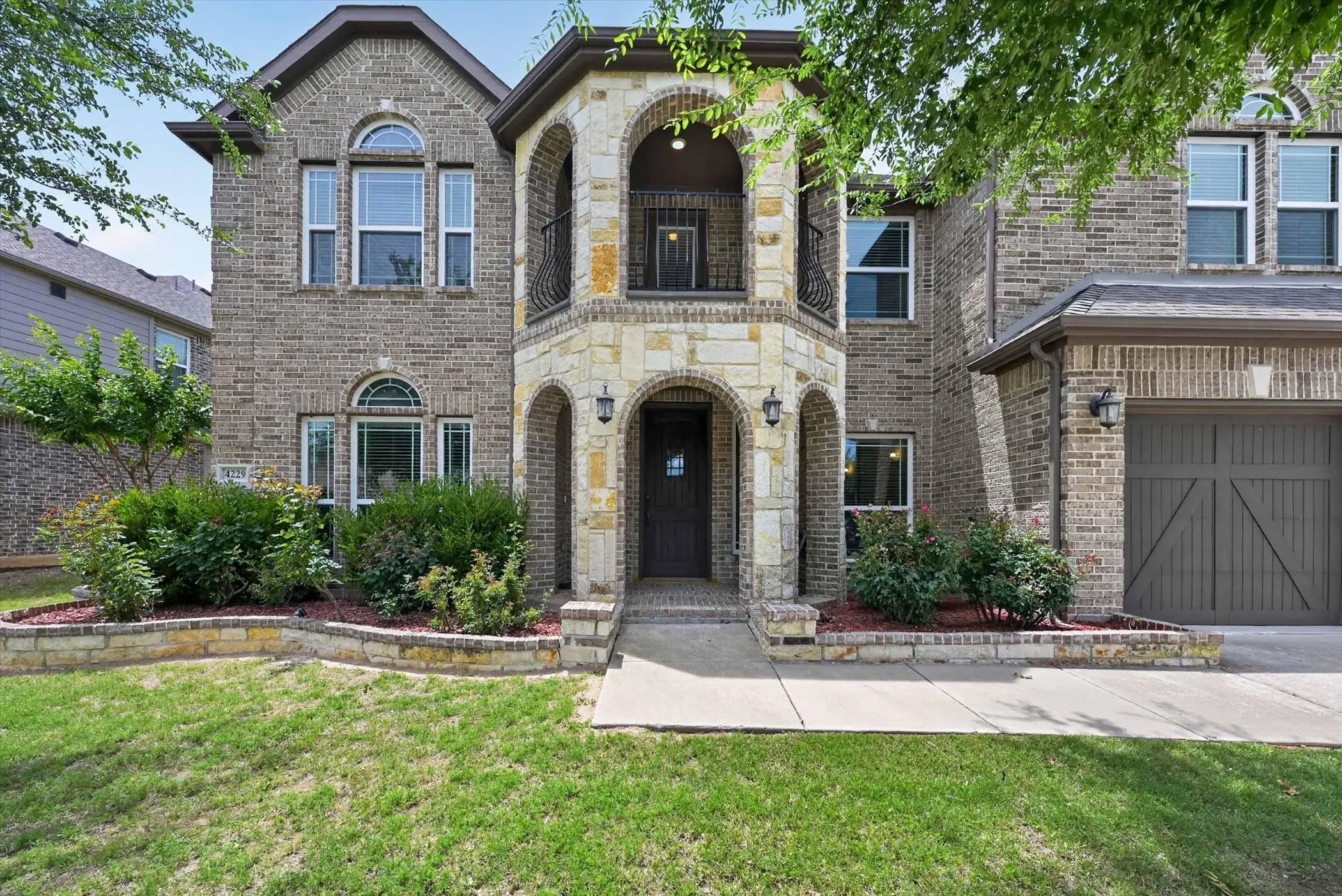 Two-story brick facade featuring a stone entryway with arched accents, a second-story balcony with wrought iron railing, an arched transom window, and a paneled garage door