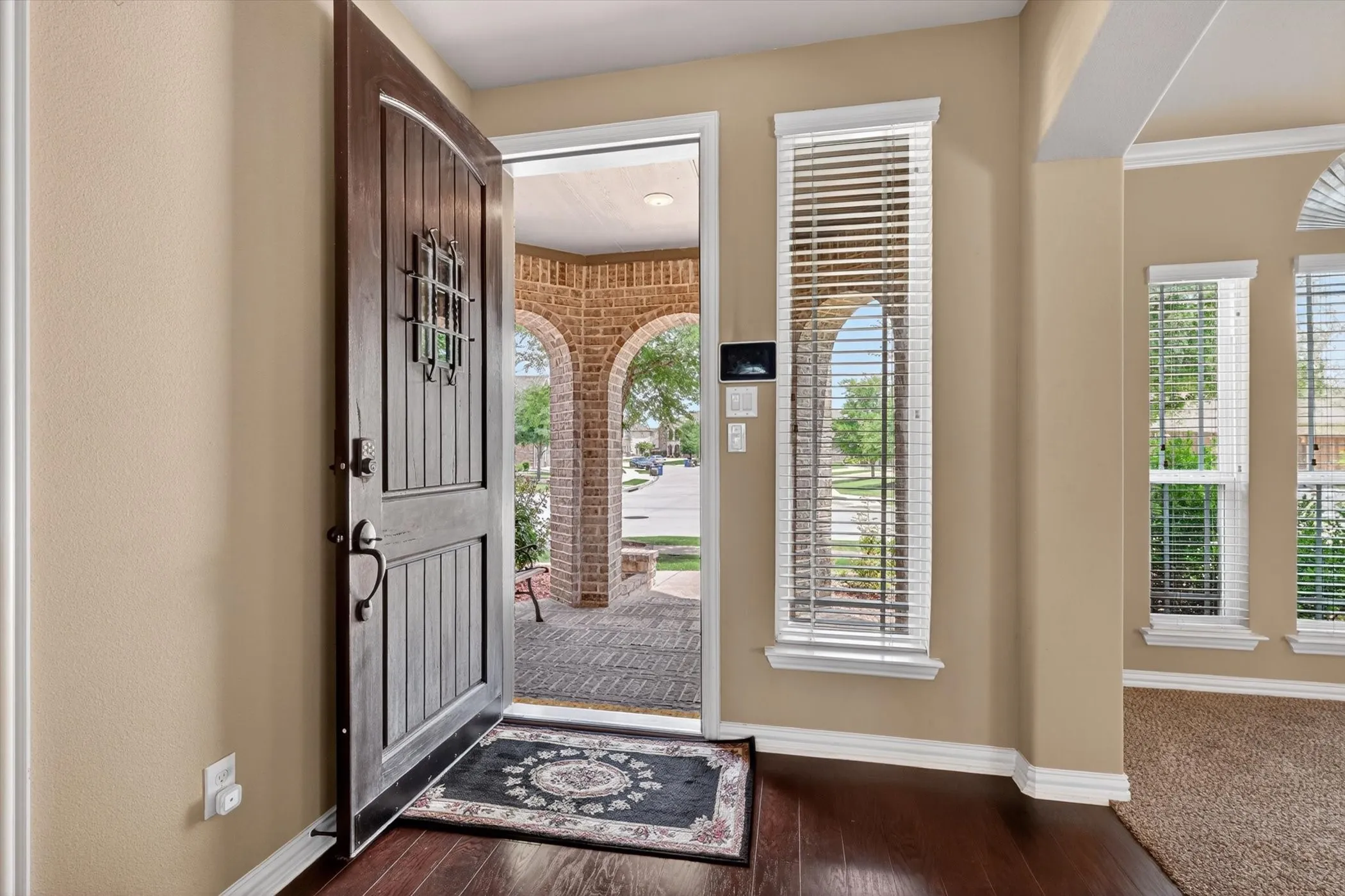 Rich wood-finish flooring in an entry foyer featuring a dark wood front door with decorative ironwork, tall sidelight windows with blinds, and light tan wall paint