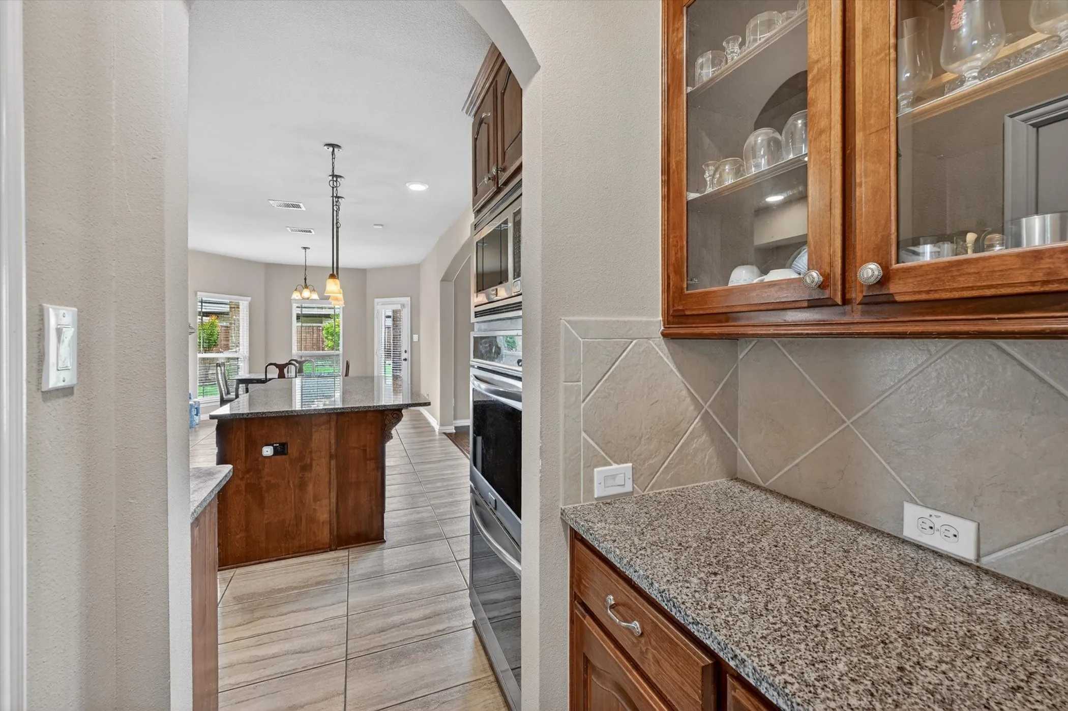 Kitchen with a large central island, granite countertops, tile flooring, built-in double ovens, and glass-front upper cabinetry