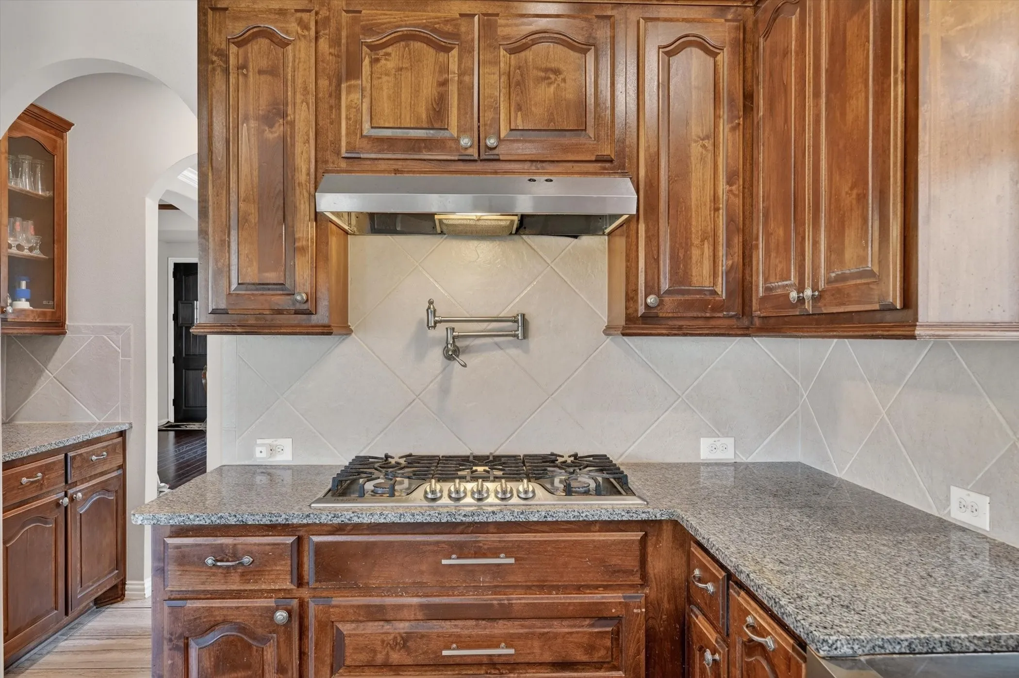 Kitchen featuring rich wood cabinetry, granite-style countertops, stainless steel gas range, coordinating range hood, and a diamond-pattern tile backsplash with a pot filler faucet