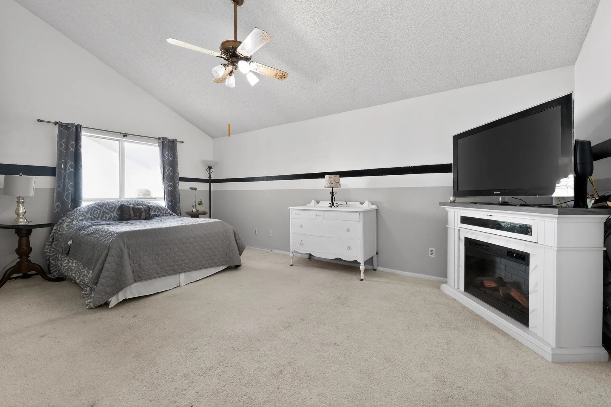 Spacious bedroom featuring vaulted ceilings, a ceiling fan, light carpeting, a large window, and an electric fireplace