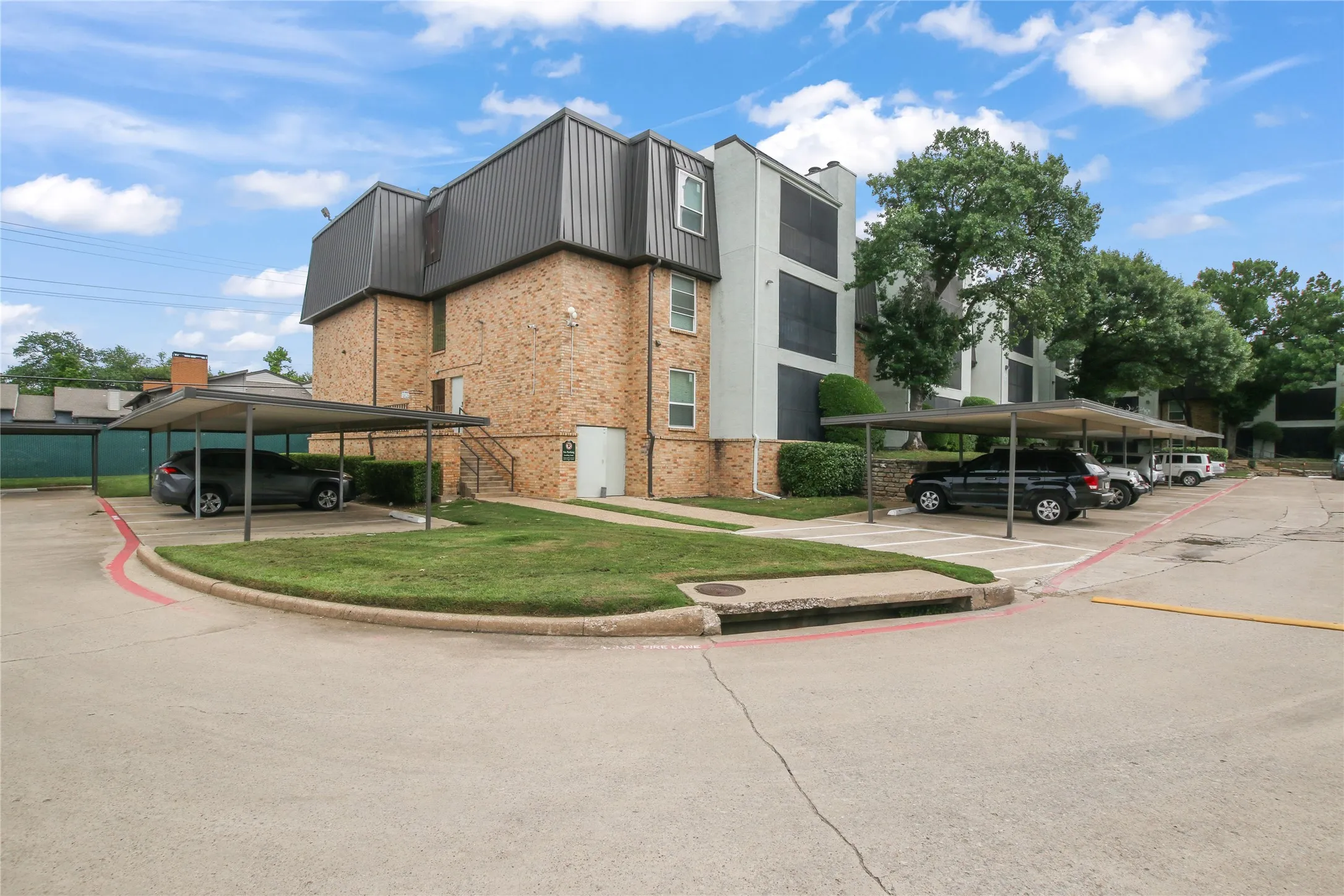 Brick exterior building with covered parking, mature trees, manicured landscaping, and concrete pathways