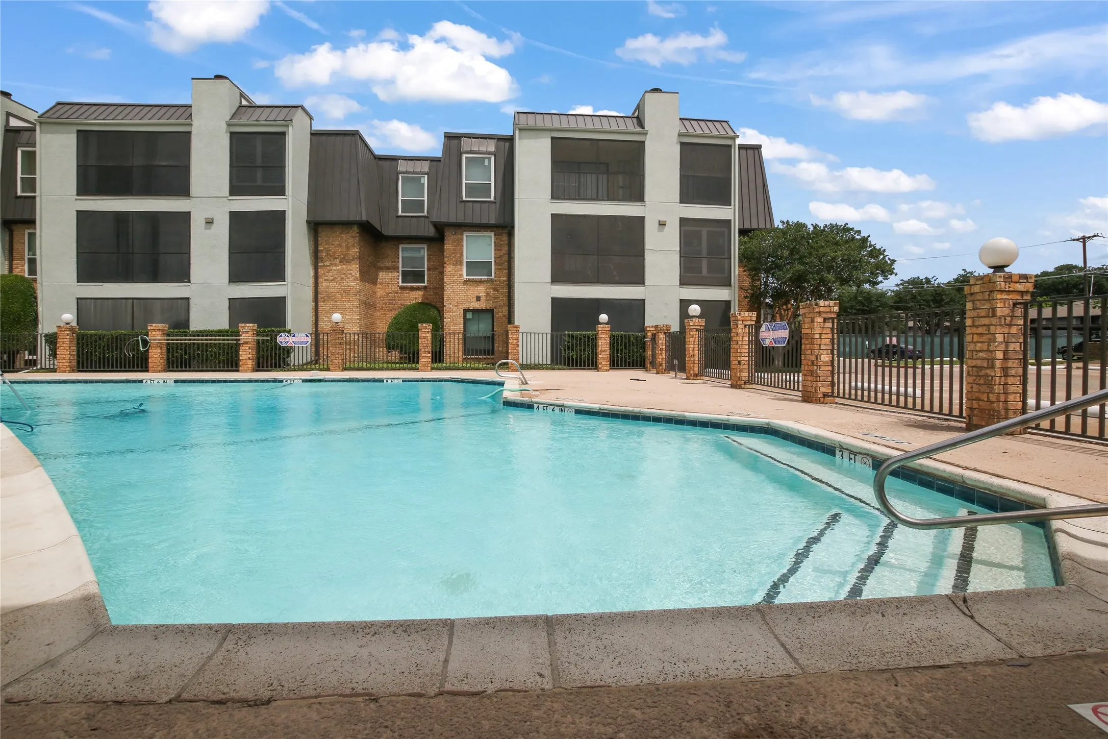 Outdoor swimming pool with integrated steps, surrounded by a concrete deck and a brick and metal fence