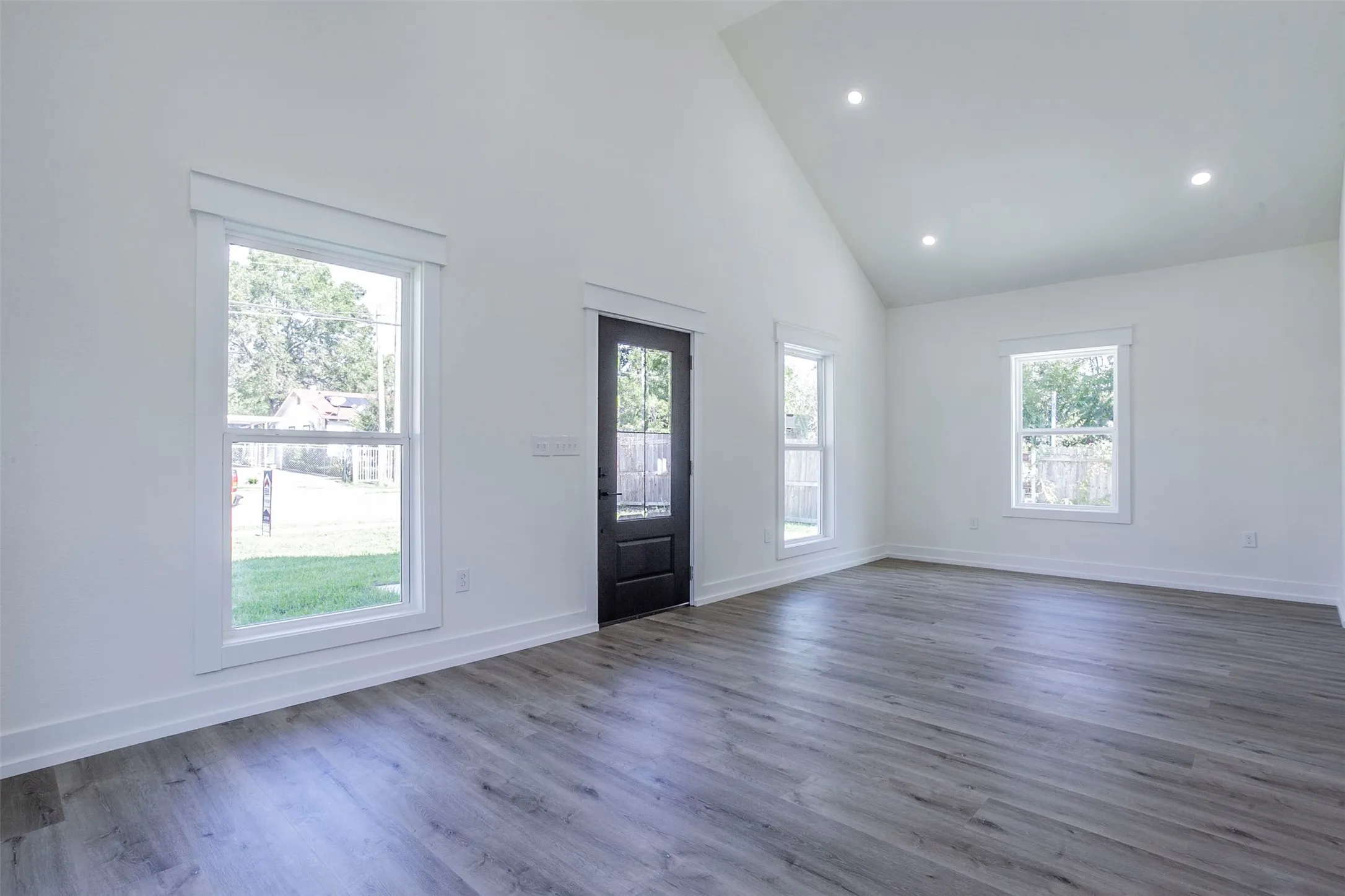 Spacious room featuring vaulted ceilings, recessed lighting, wood-finish flooring, white trim, and a dark paneled door with glass inserts