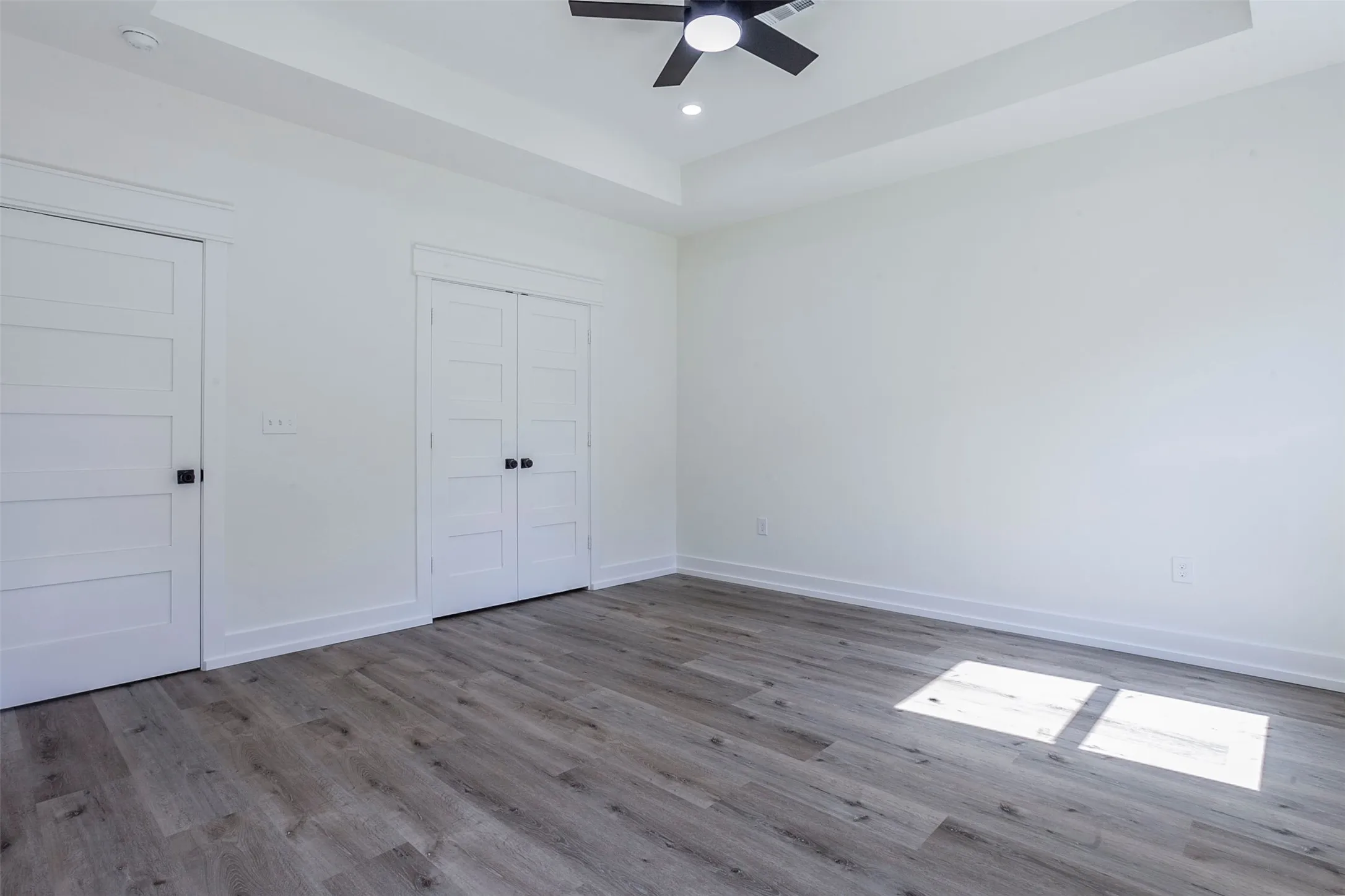Spacious room featuring wood-finish flooring, white walls, and a tray ceiling with a built-in ceiling fan