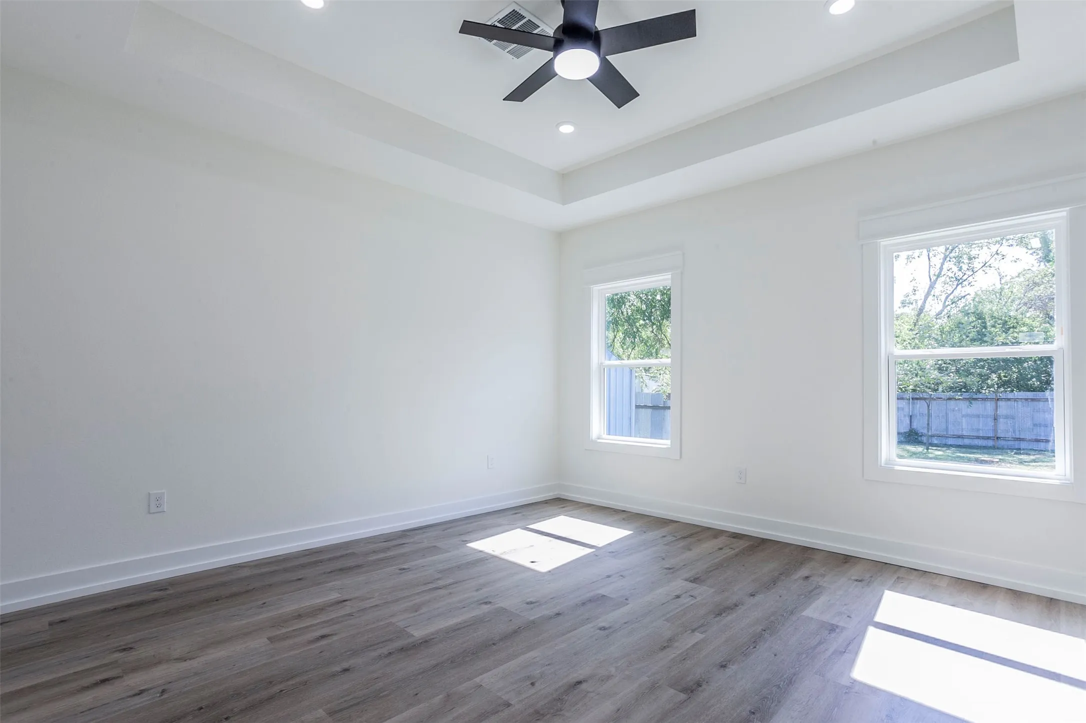 Bright room featuring wood-finish flooring, two large windows, a tray ceiling, recessed lighting, and a ceiling fan