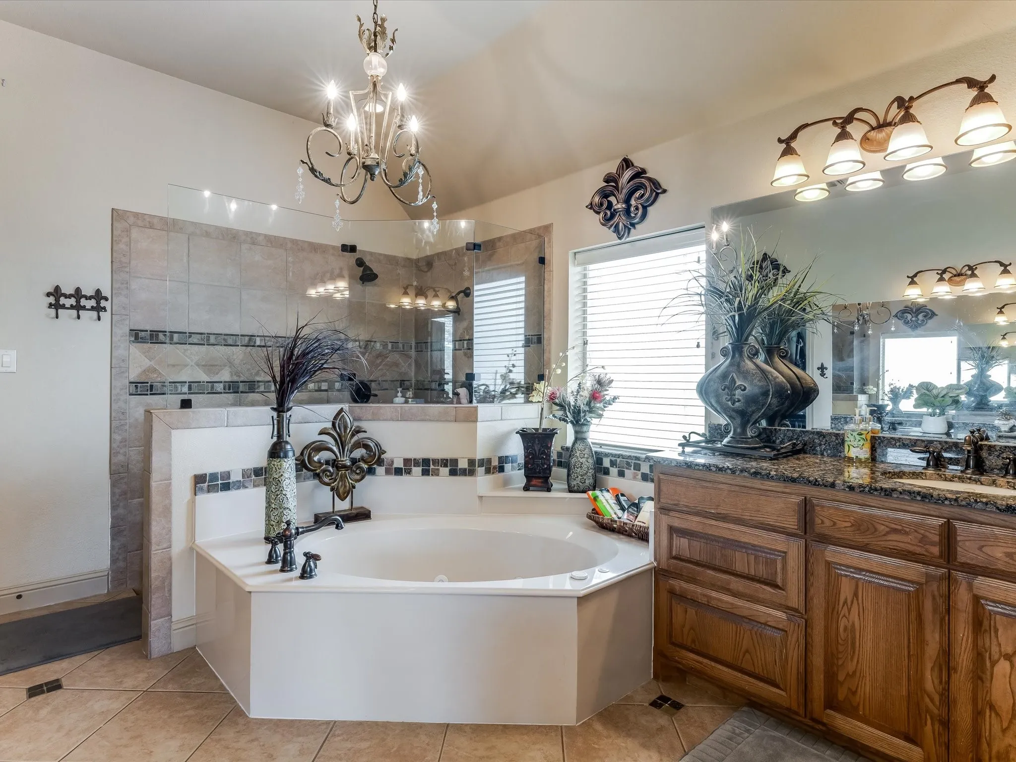 Tiled bathroom featuring a corner soaking tub, glass-enclosed shower, a double vanity with stone-finish countertop, and decorative chandelier lighting