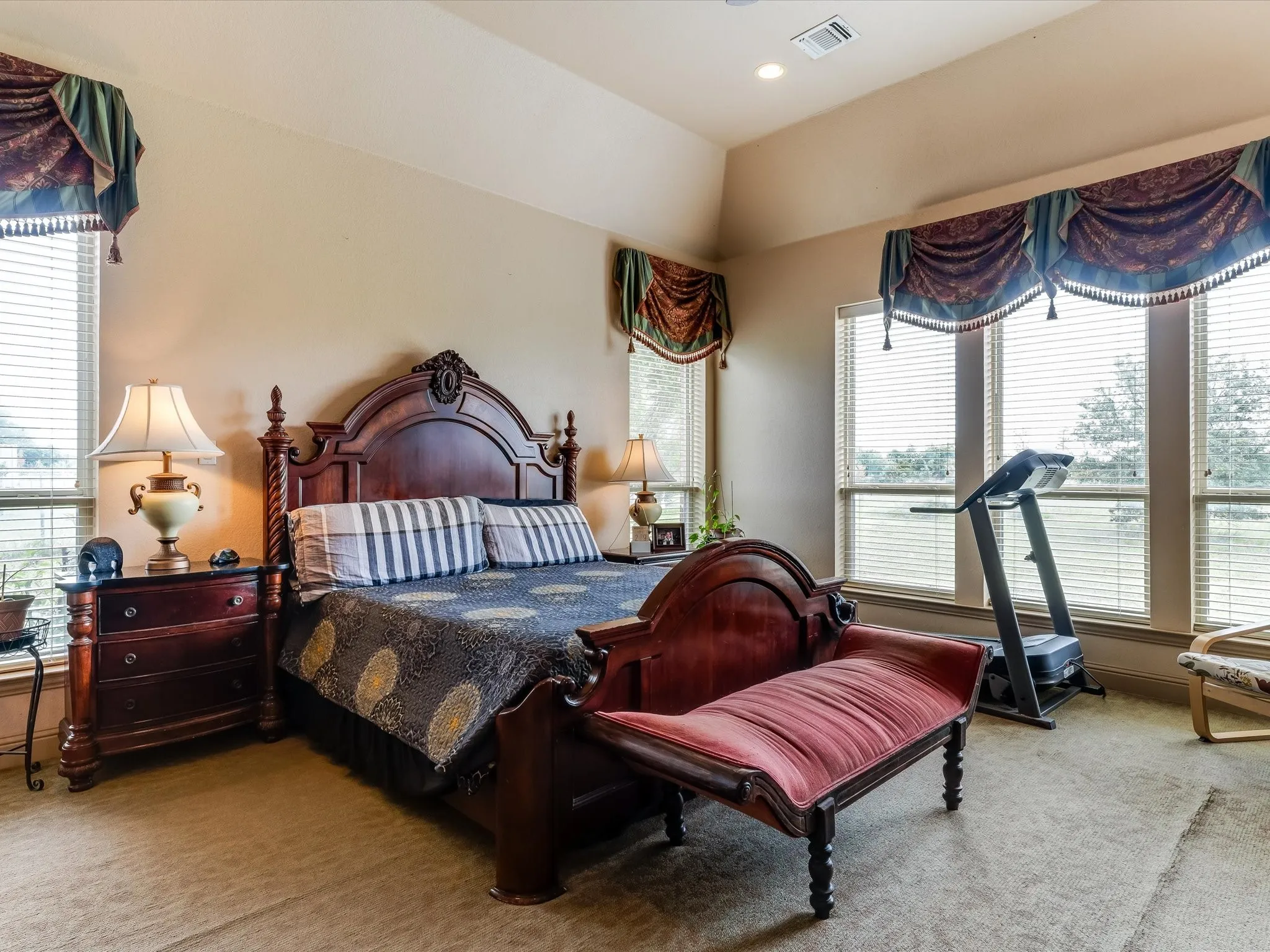 Spacious bedroom featuring multiple large windows with horizontal blinds, decorative valances, and a tray ceiling