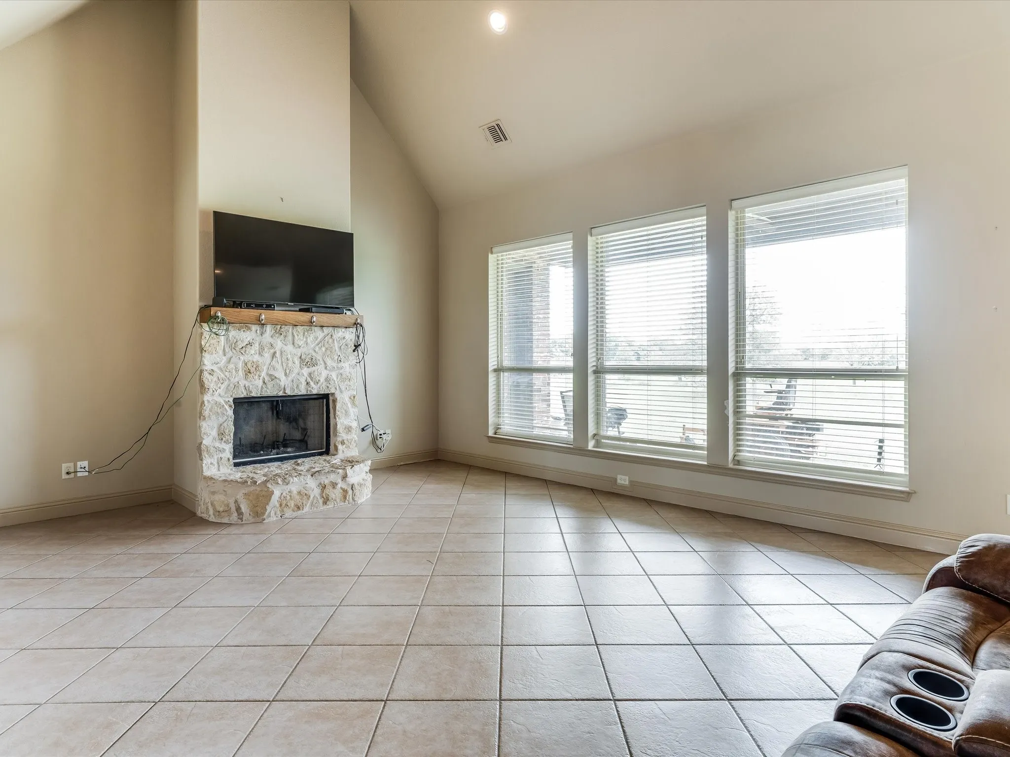 Living area featuring a floor-to-ceiling stone-clad fireplace with a hearth, tile flooring, vaulted ceiling, and three large windows with horizontal blinds