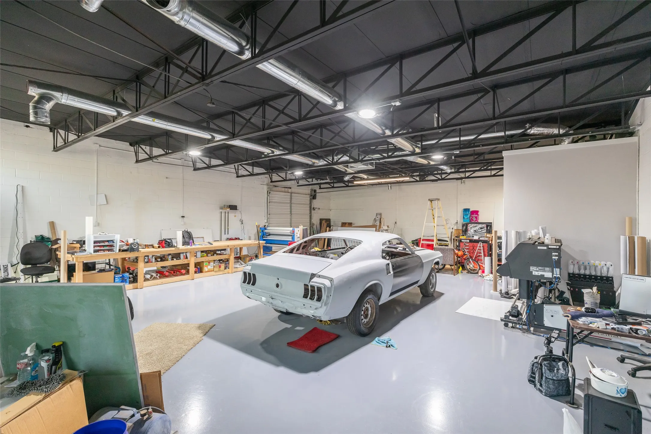 Spacious garage workshop featuring a large overhead door, exposed ductwork, multiple fluorescent light fixtures, a high ceiling with exposed structural beams, and a light gray epoxy floor