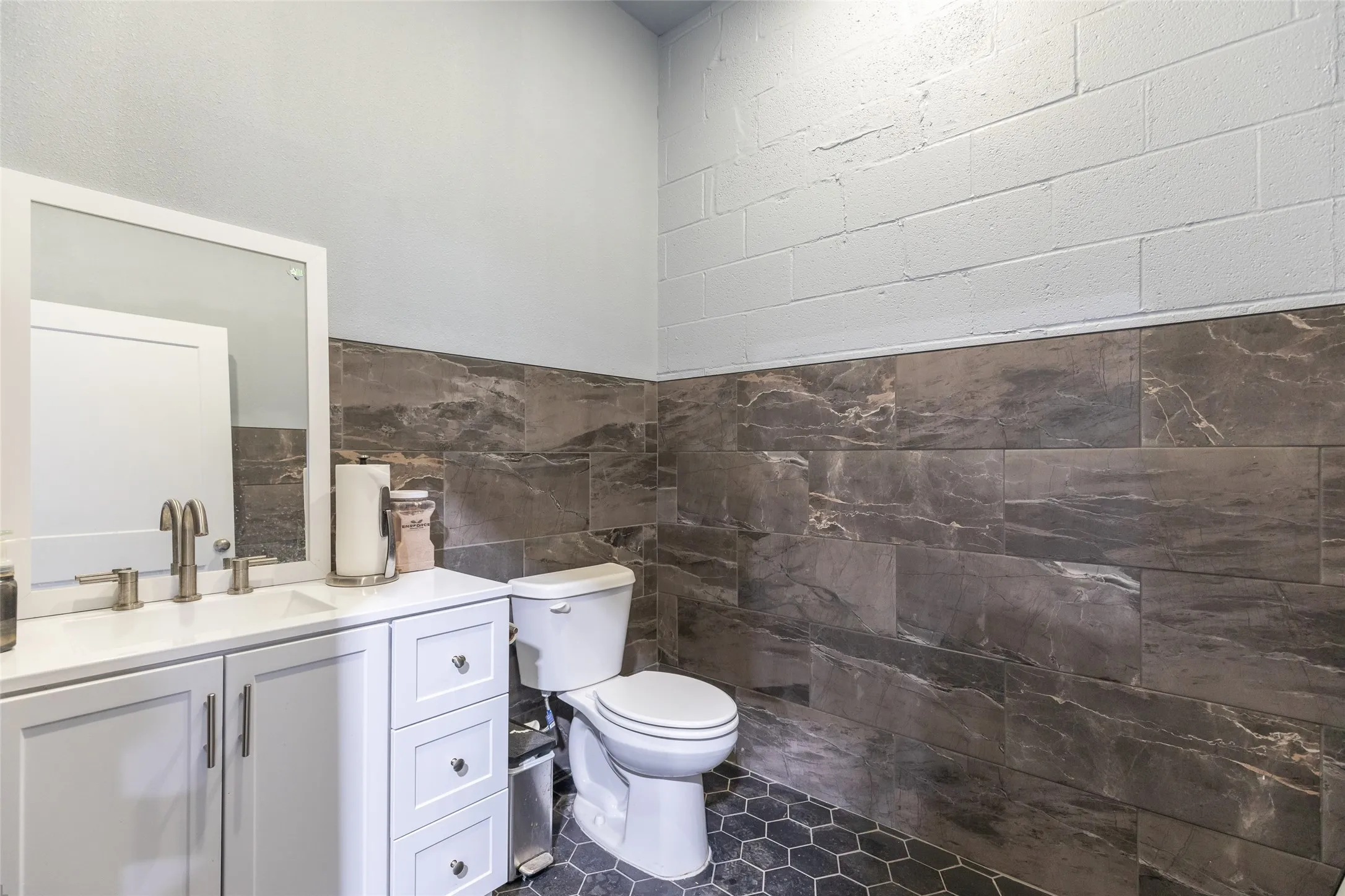 Contemporary bathroom featuring a white vanity with integrated sink, brushed nickel faucet, large rectangular mirror, a white toilet, and dark hexagon floor tiles