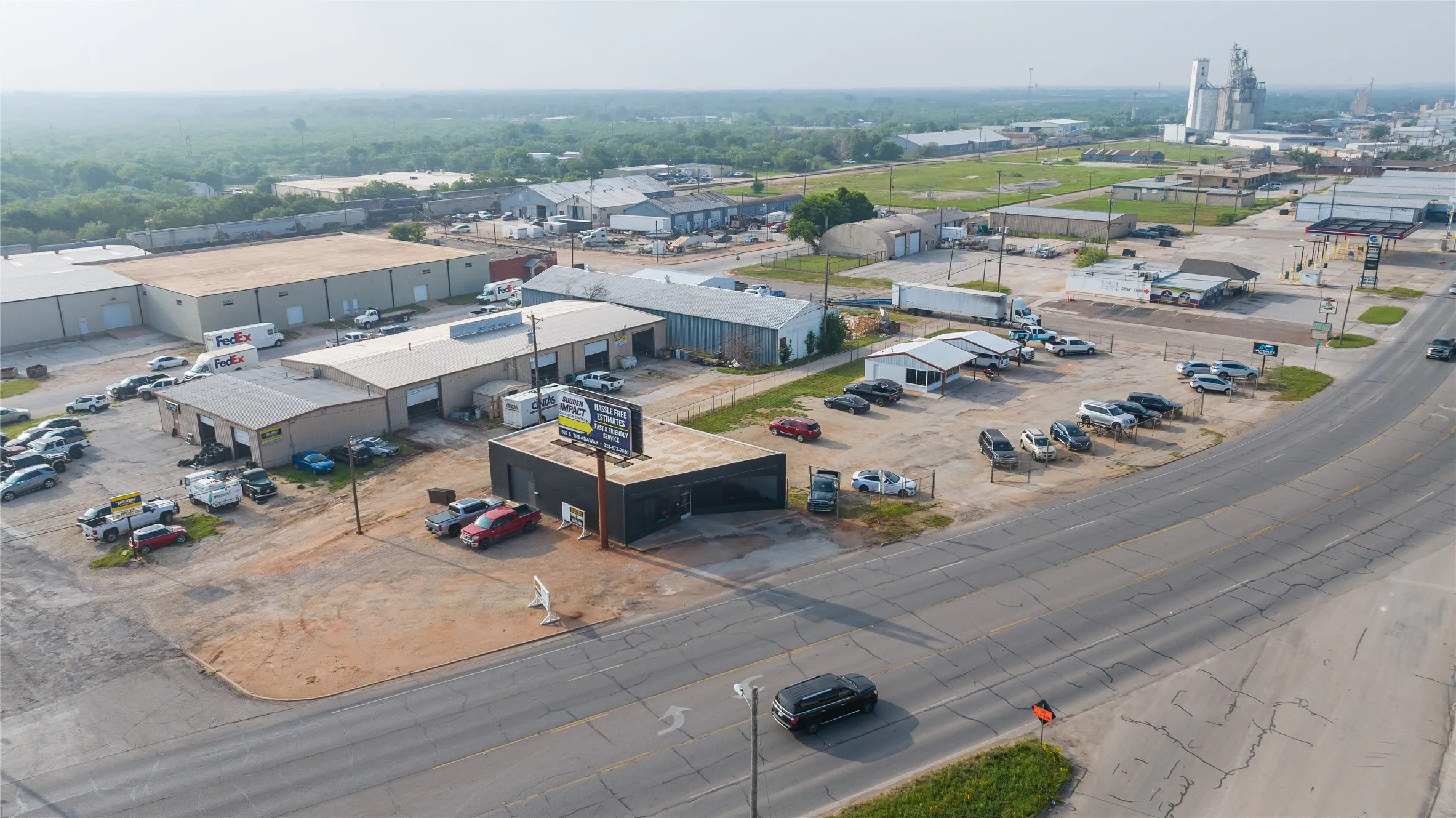 Commercial property featuring multiple single-story buildings with roll-up doors, extensive paved and unpaved parking areas, and roadside visibility