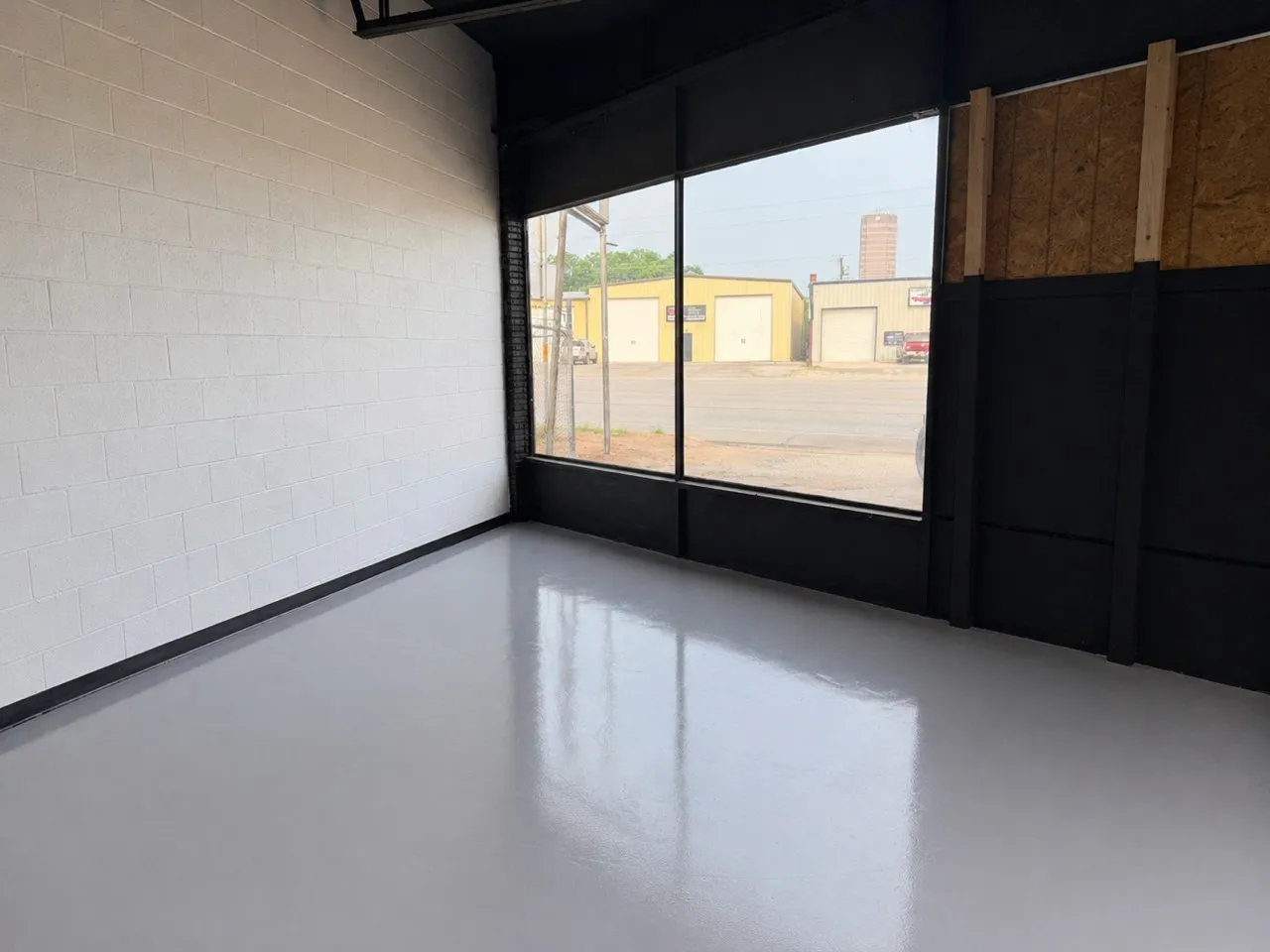 Open interior space featuring a large storefront window, light gray polished concrete flooring, white painted cinder block wall, black painted baseboards, and black exposed ceiling beams