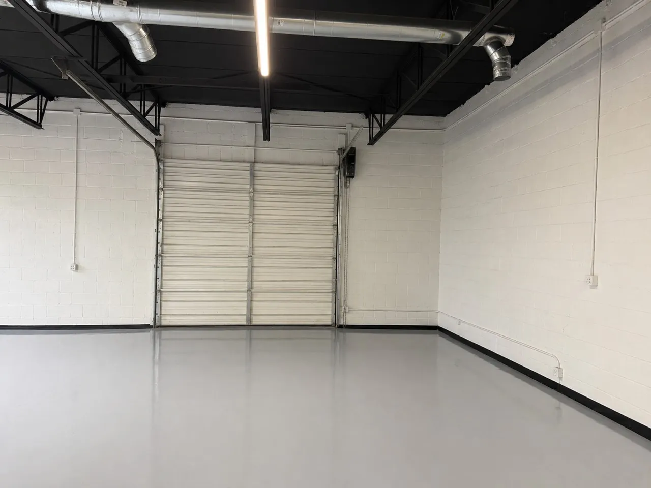 Spacious industrial interior featuring a large roll-up door, polished concrete flooring, exposed ceiling trusses, overhead ductwork, and fluorescent lighting