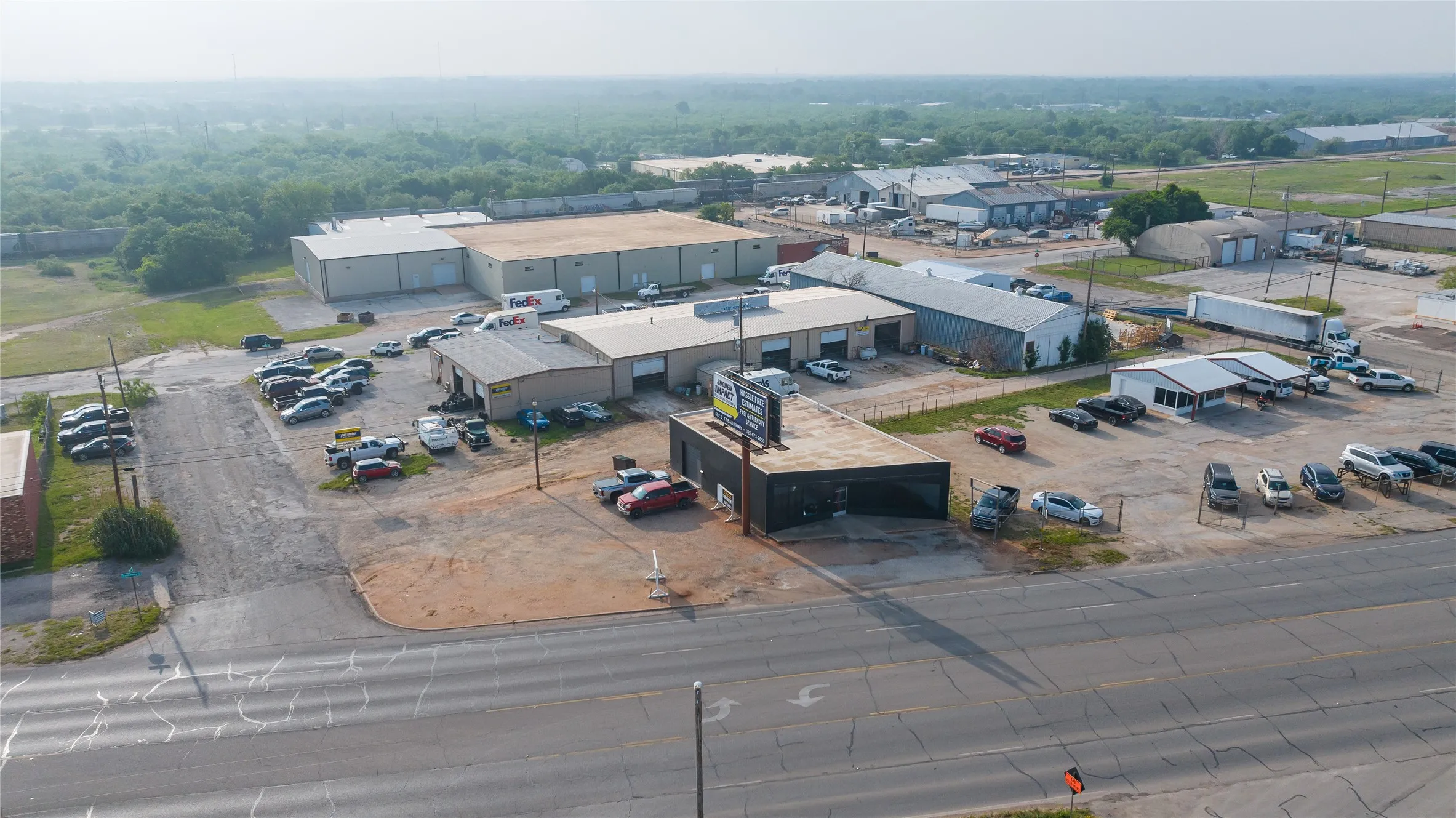 Aerial perspective highlighting a commercial property with multiple industrial-style buildings, extensive paved and gravel parking areas, and a prominent roadside building