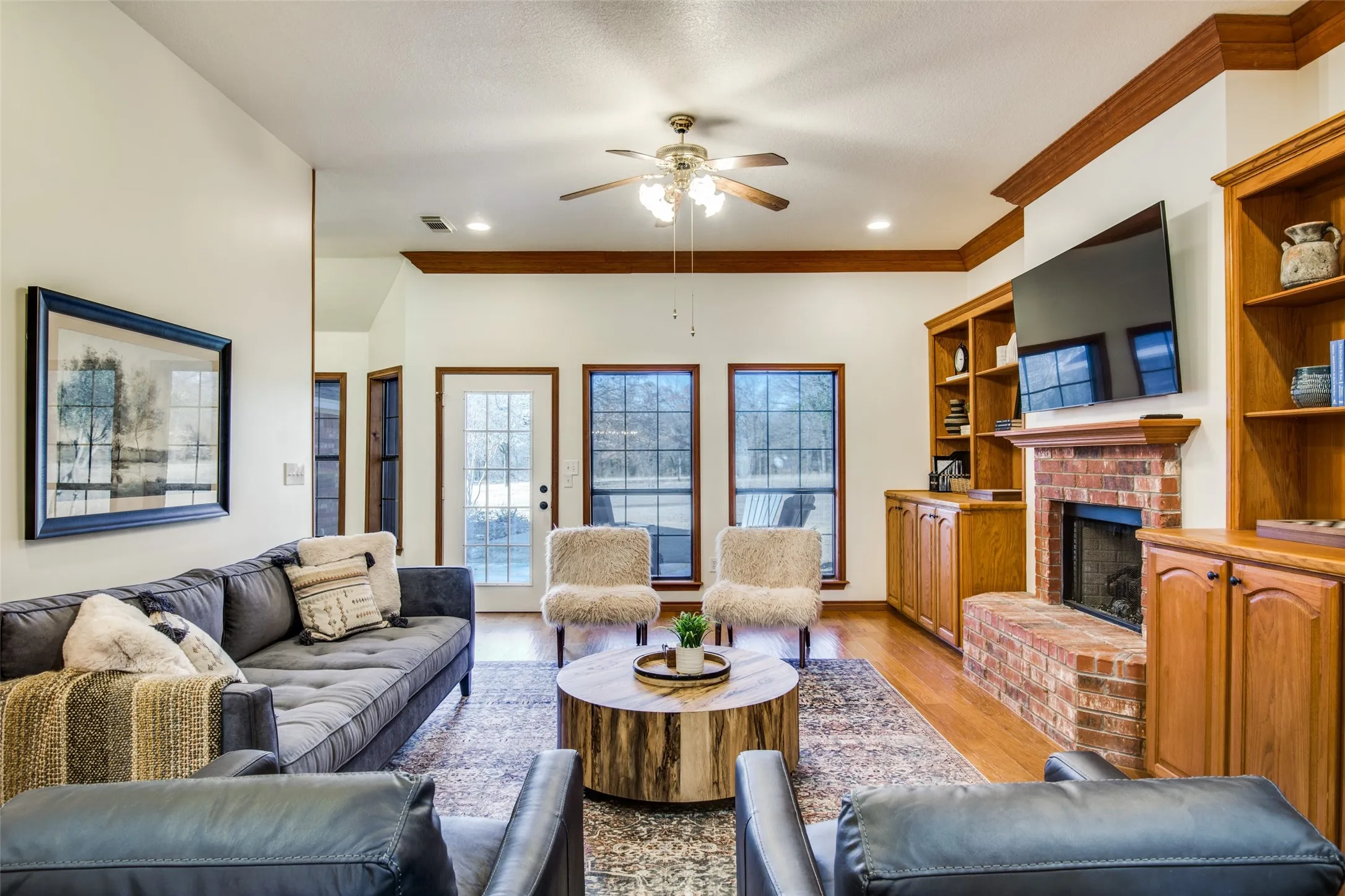 Bright living area with wood-finish flooring, a brick fireplace with wood mantel, and built-in wood cabinetry and shelving