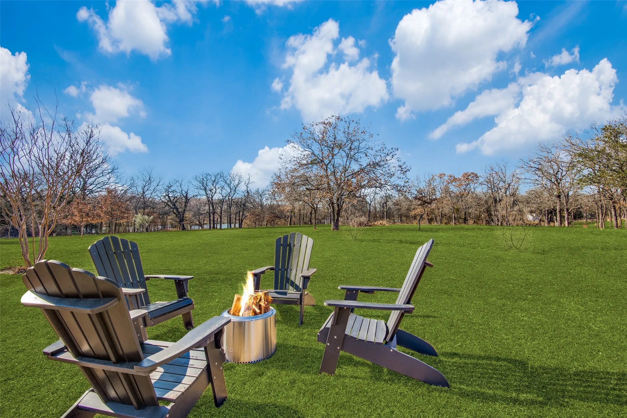 Expansive green lawn with a fire pit surrounded by Adirondack chairs, bordered by mature trees