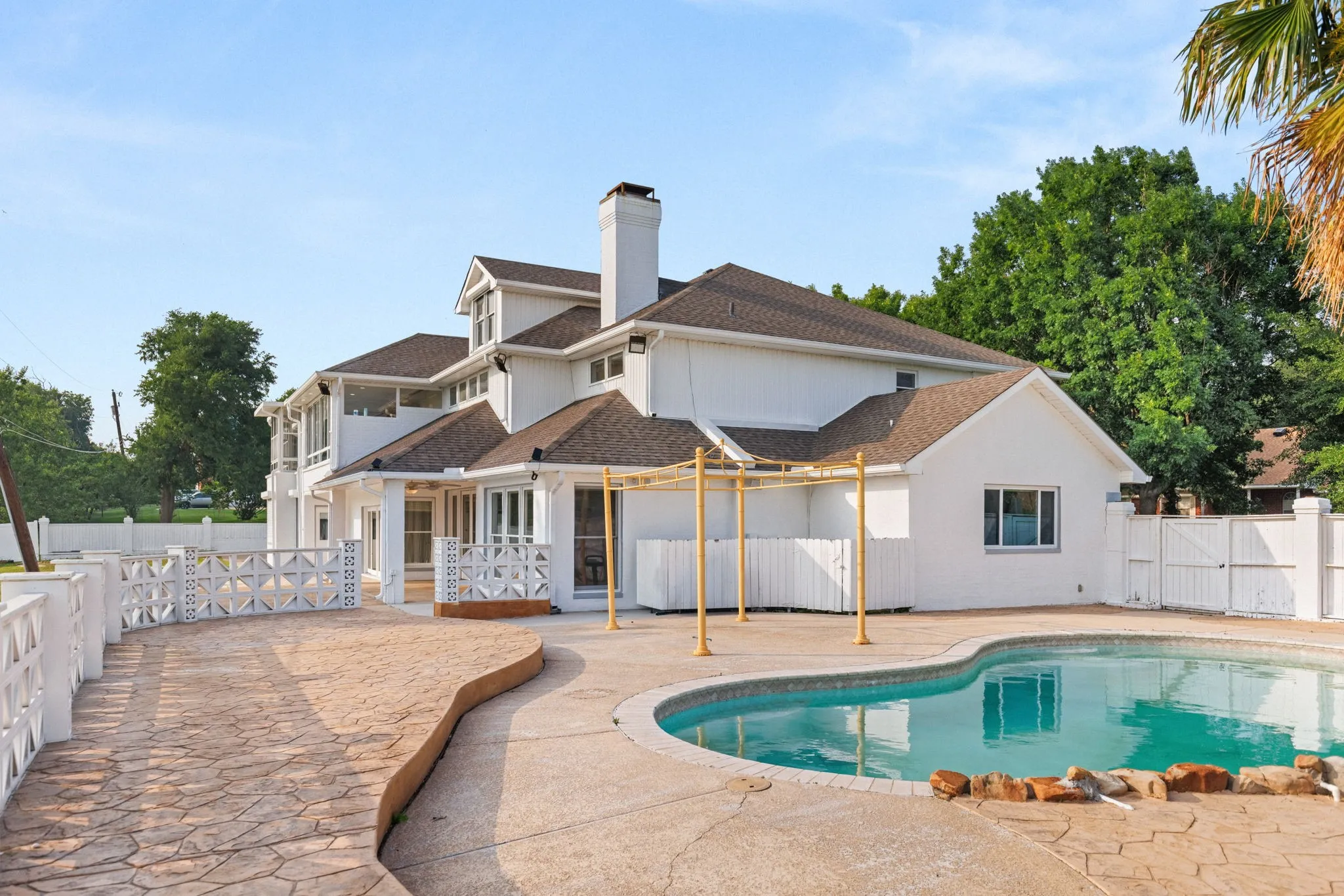 Rear exterior featuring a swimming pool, stamped concrete decking, white painted brick, a white privacy fence, and a decorative concrete block wall