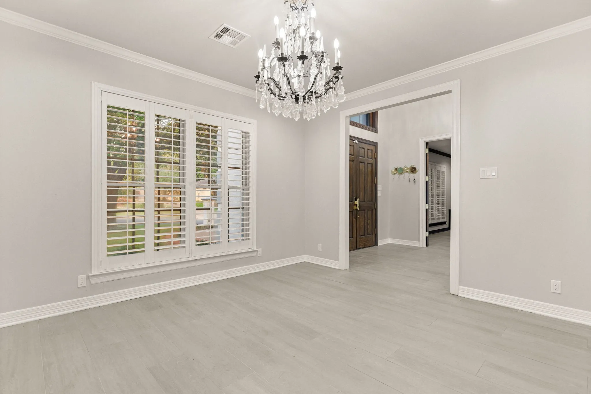 Bright interior space featuring a large window with white plantation shutters, a multi-tiered chandelier, light-toned wood-finish flooring, and white crown molding