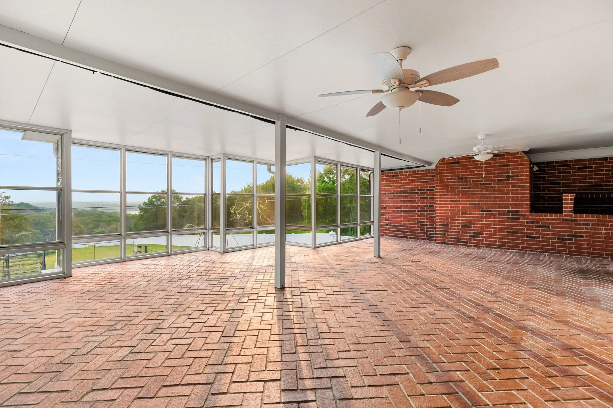 Expansive enclosed patio featuring herringbone brick flooring, floor-to-ceiling windows, two ceiling fans, and a built-in brick grilling area
