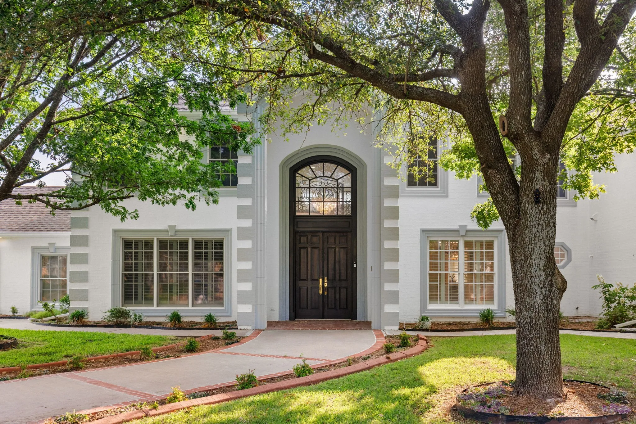 Stately exterior featuring a grand arched entry door with decorative ironwork, a curved paved walkway, mature trees, and landscaping elements