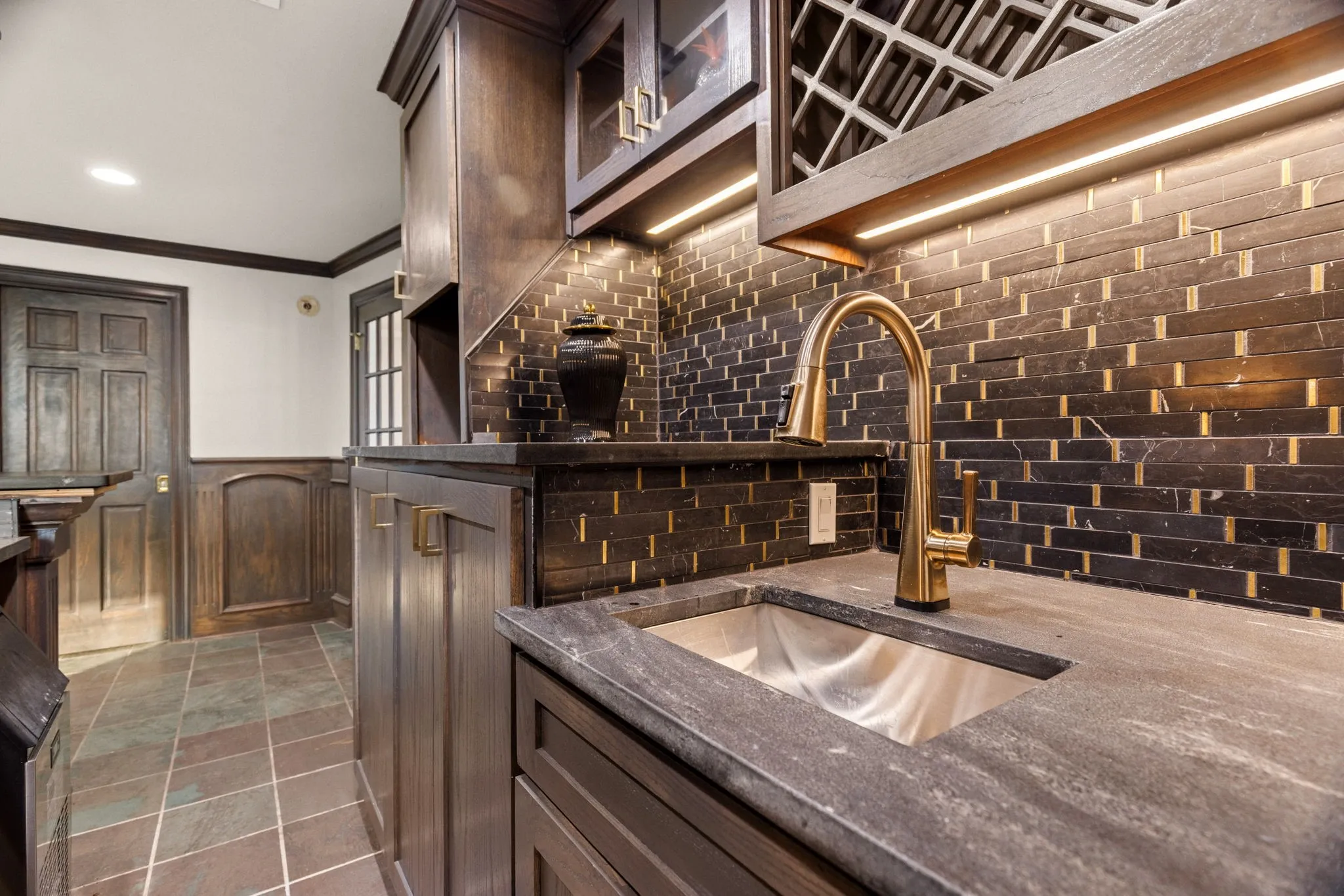 Wet bar featuring a gold-tone faucet, undermount sink, stone countertop, and dark wood cabinetry with under-cabinet lighting