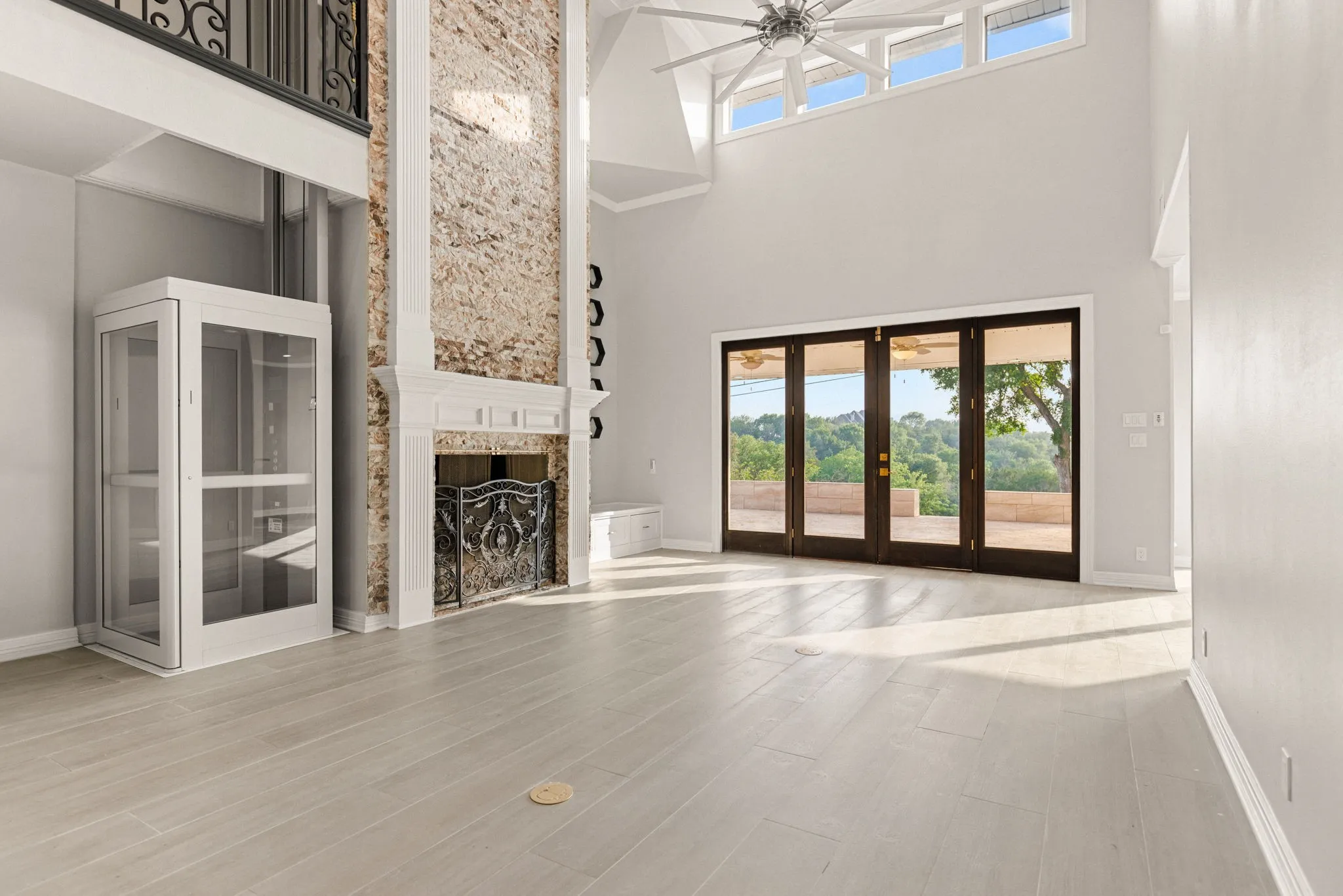 Bright, open living space featuring a two-story stone fireplace with ornate ironwork, a glass-enclosed interior lift, and expansive folding glass doors opening to outdoor scenery