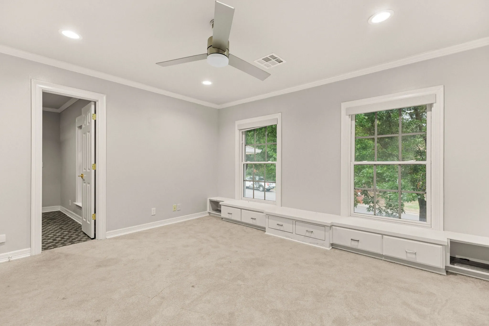 Spacious interior room featuring light gray wall paint, crown molding, a ceiling fan, recessed lighting, and two windows with built-in storage benches