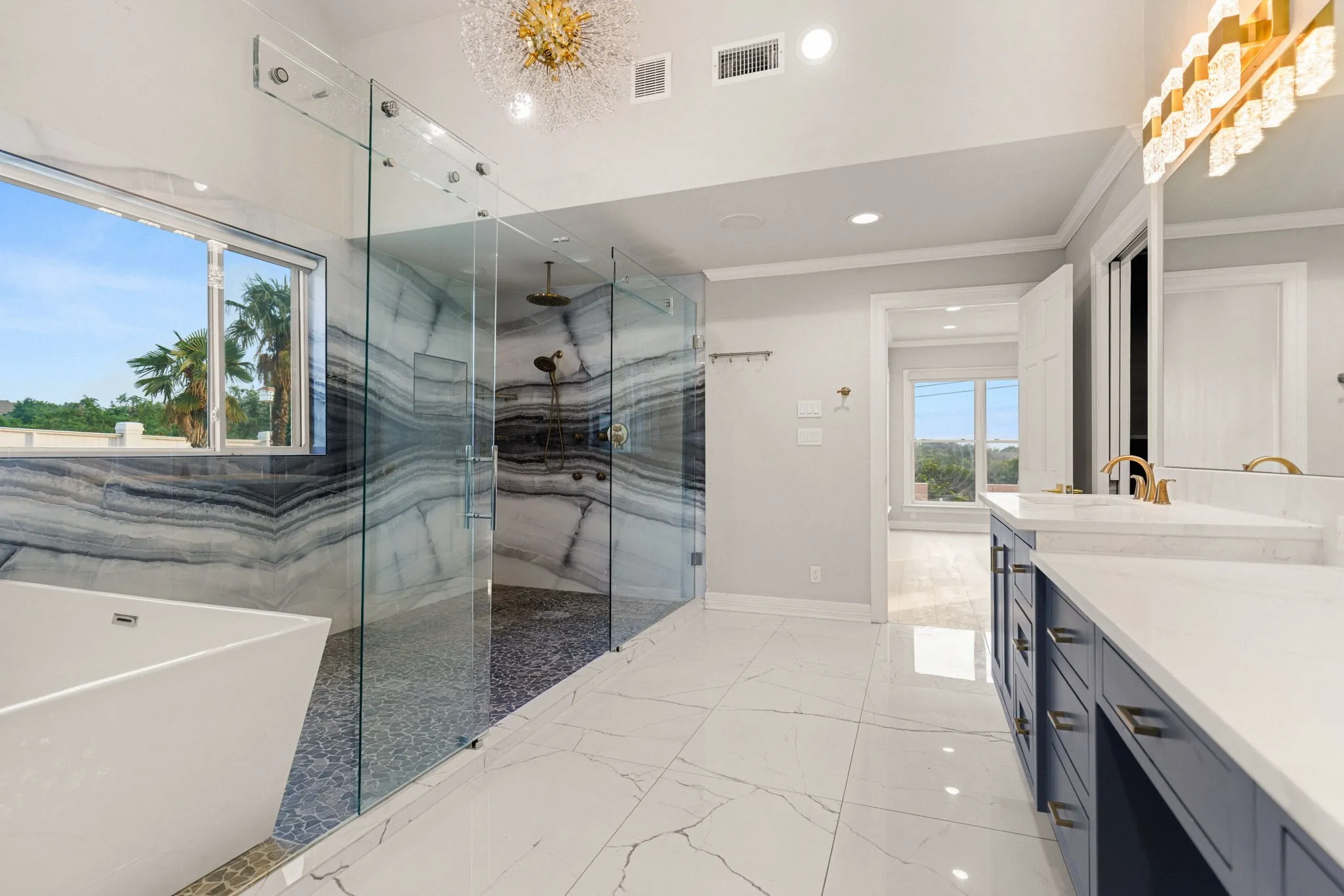 Spa-inspired bathroom featuring a freestanding soaking tub, a large walk-in shower with patterned stone tile and glass enclosure, a double vanity with white countertops and gold-finish fixtures, and large-format tile flooring
