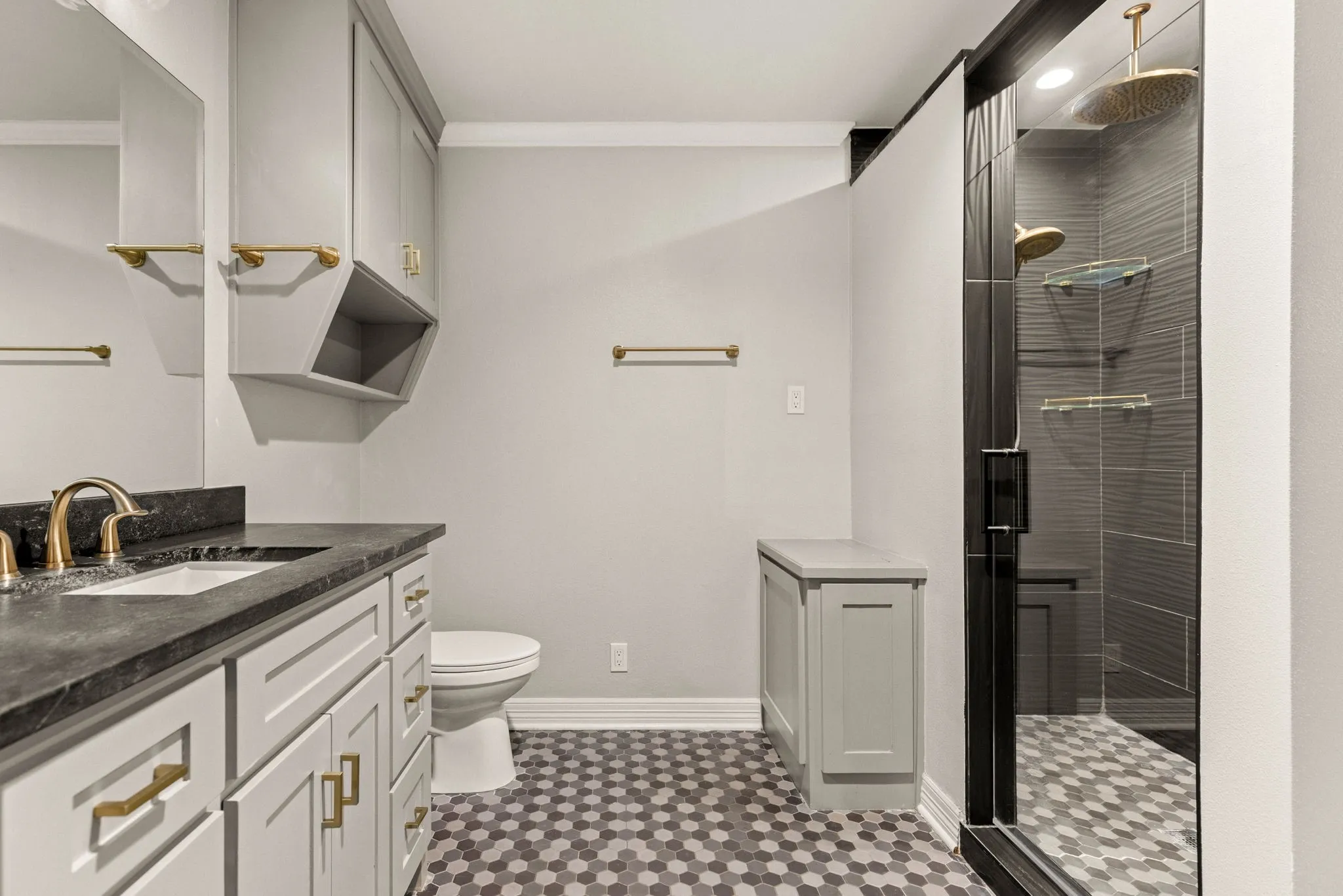 Bathroom featuring hexagon tile flooring, a walk-in shower with glass enclosure, a vanity with a dark countertop and undermount sink, and light-toned cabinetry with gold-finish hardware