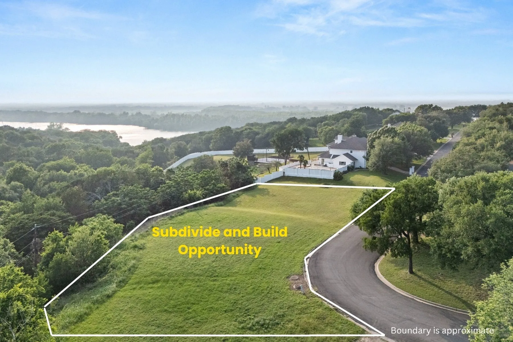 Expansive undeveloped lot featuring a paved road frontage, surrounding mature trees, and a distant water body