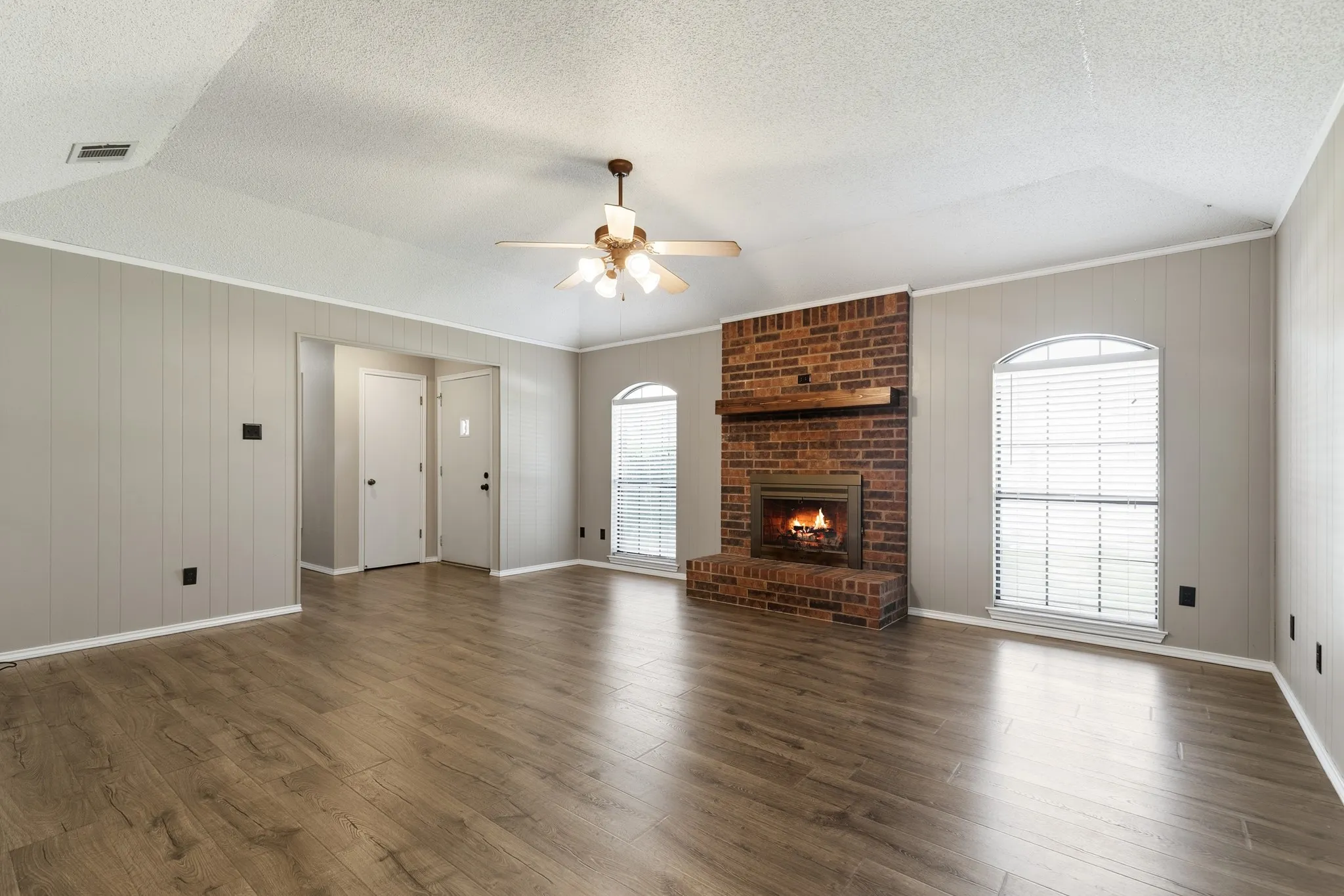 Spacious room featuring wood-finish flooring, a brick fireplace with a wood mantel, and two arched windows with blinds